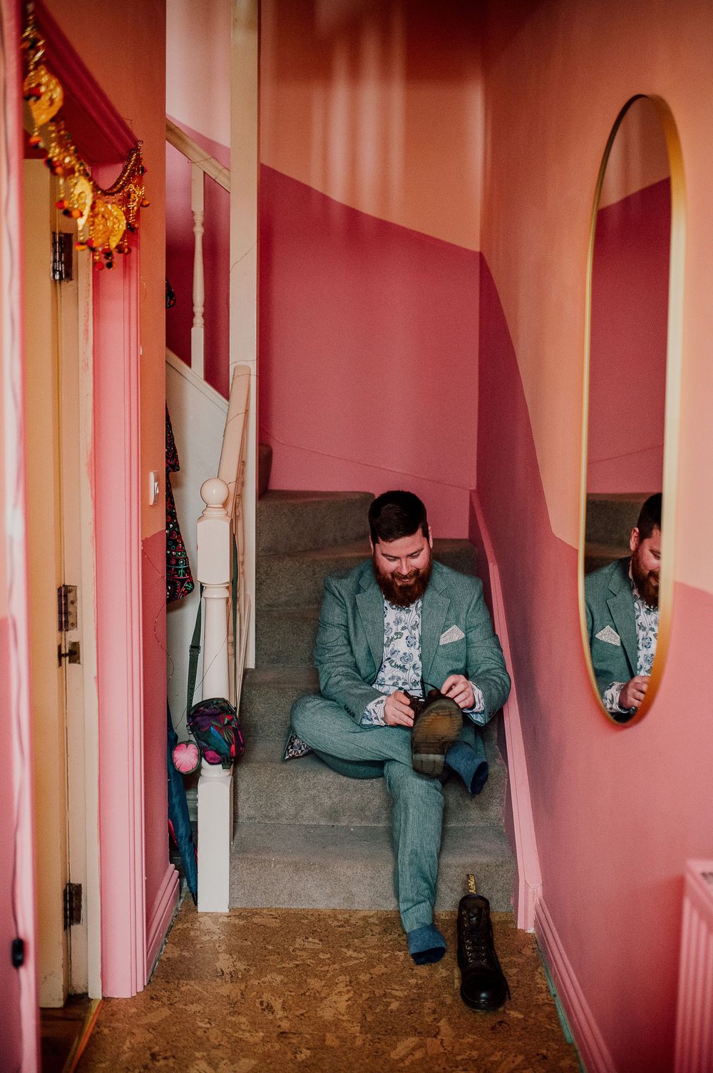 A person in a mint green suit sits on pink-walled stairs looking at their phone in a vintage-style interior.