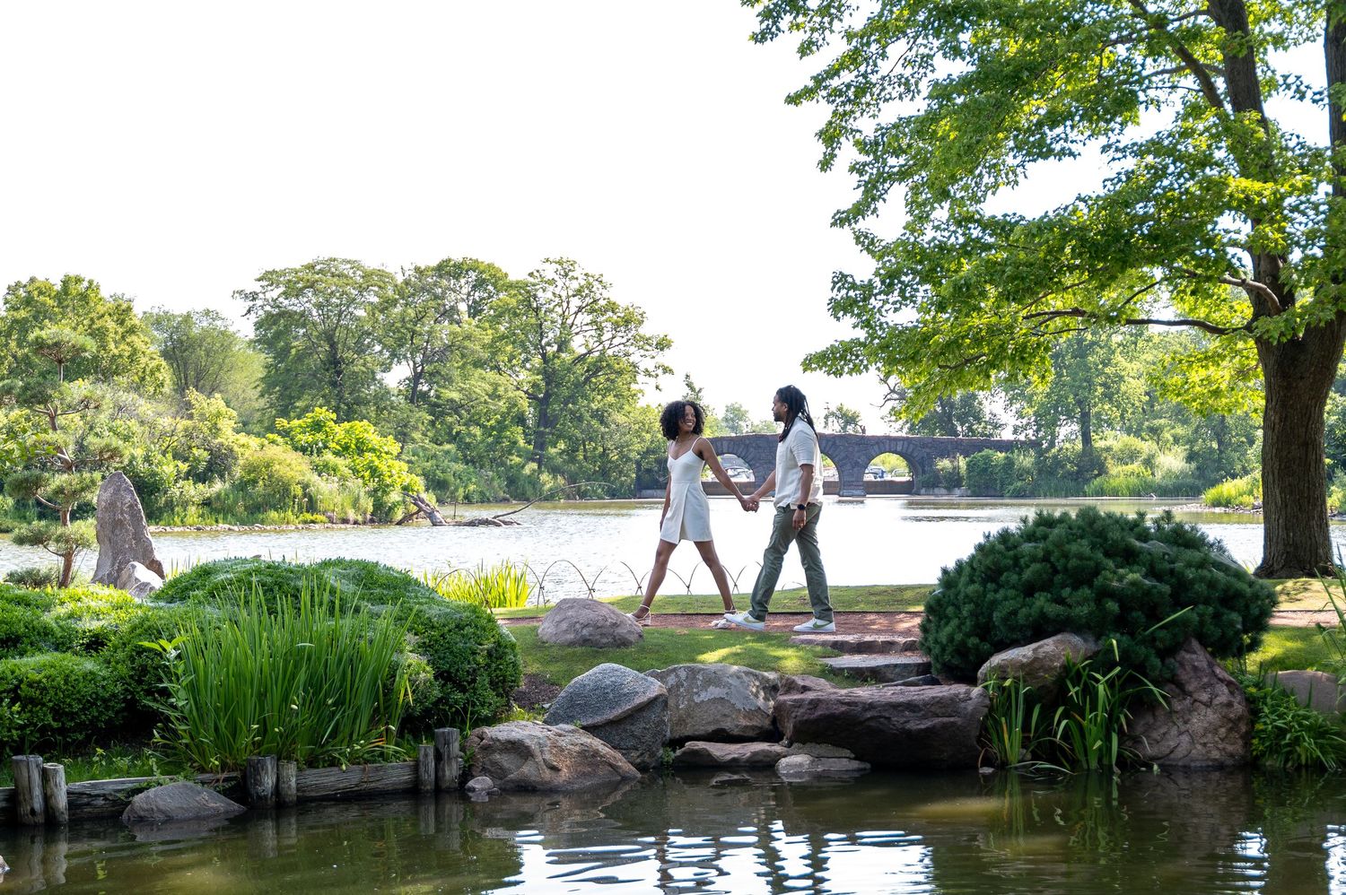 Two people walk along a scenic garden path beside a tranquil pond with rocks and greenery on a sunny summer day.