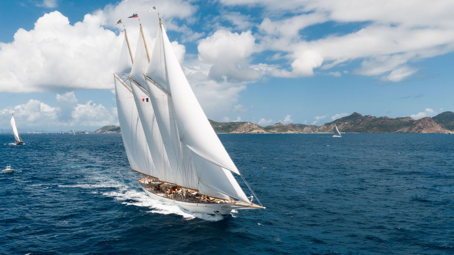A majestic sailing yacht with white sails glides across blue ocean waters under a bright cloudy sky.