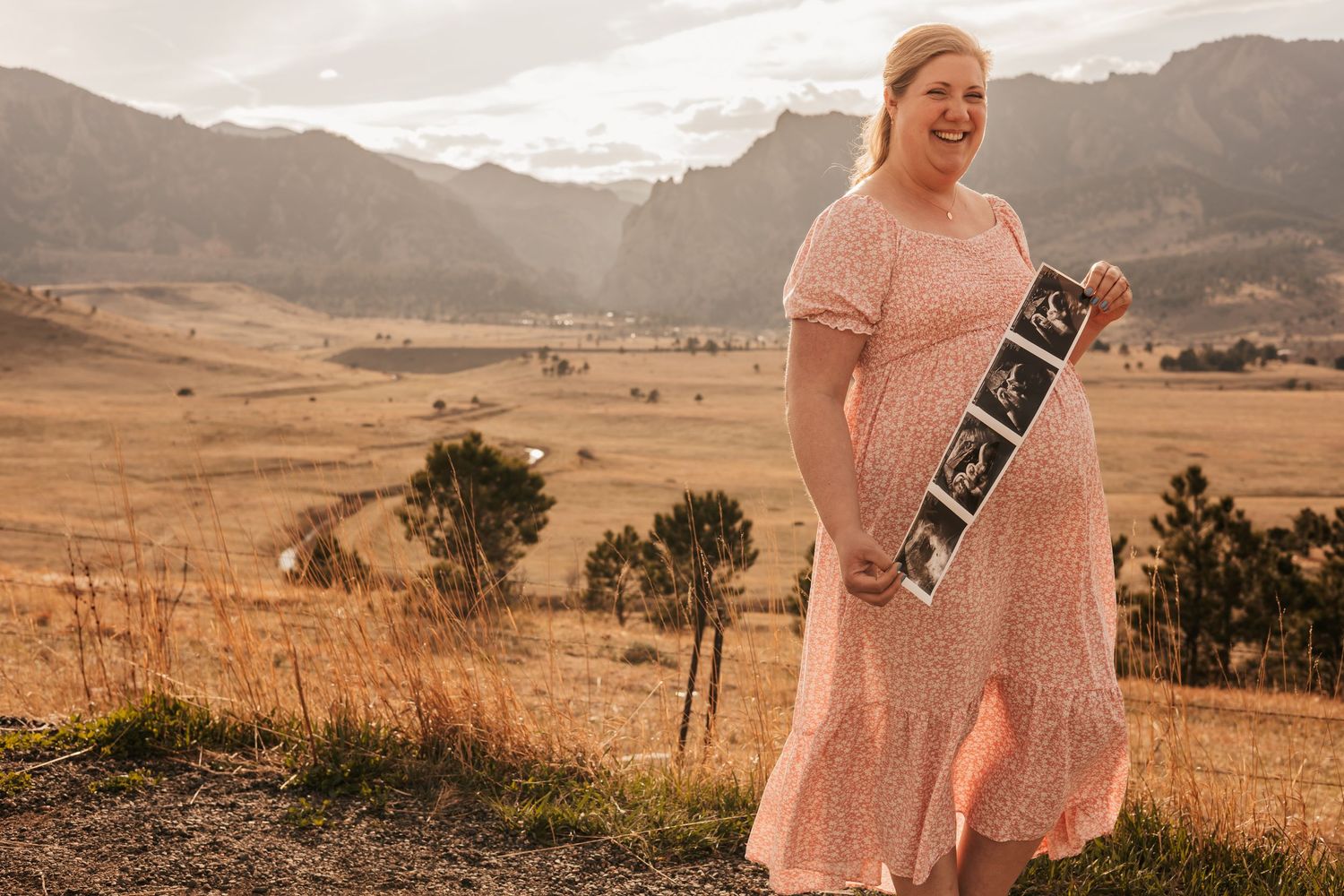 Pregnant woman in pink dress holding ultrasound photos, smiling in scenic mountain valley landscape | Maternity Photos Boulder CO