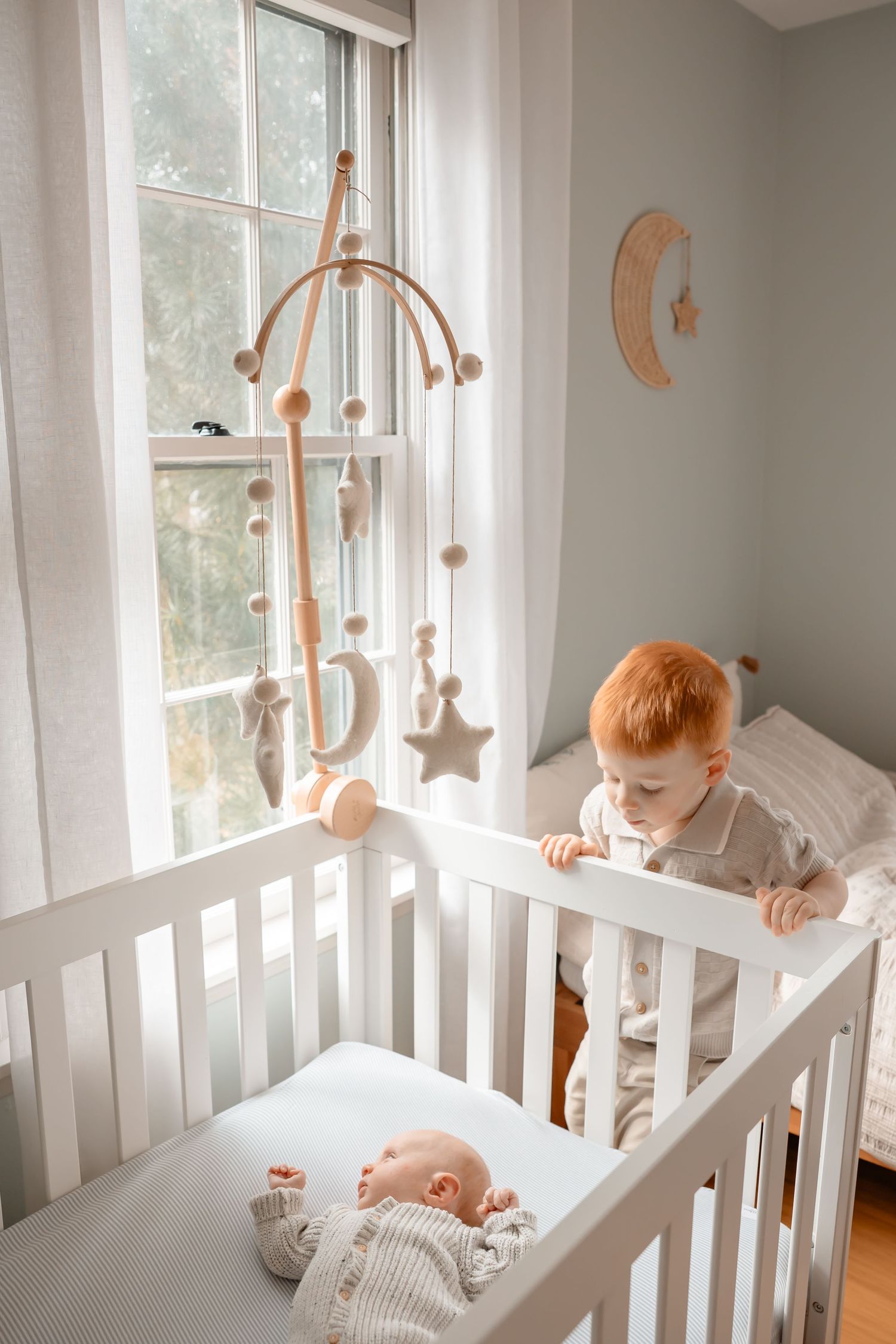 Two babies interact through white crib slats in a bright nursery with natural light and wooden mobile decorations.