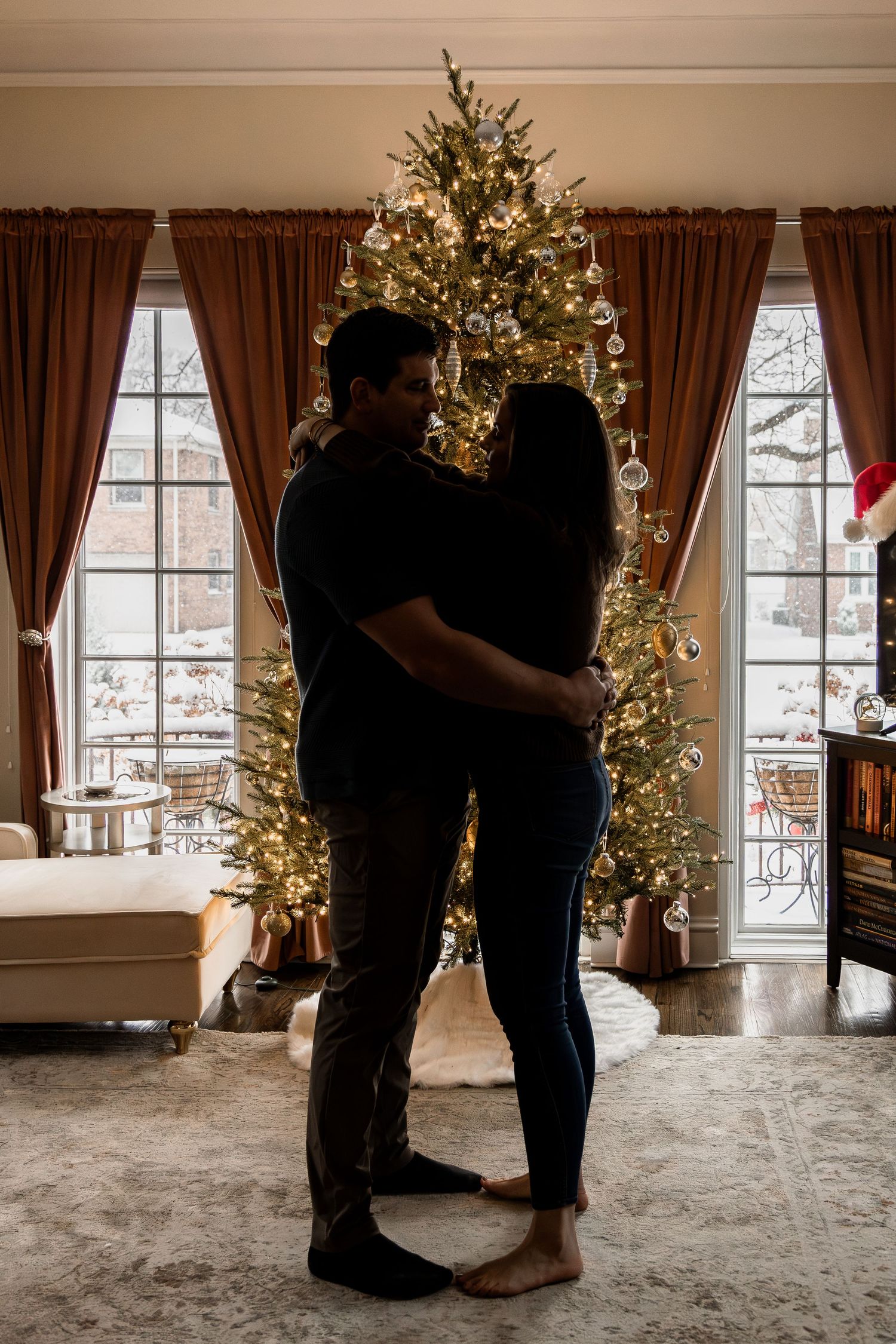 Silhouettes of a couple dancing in a romantic room with a lit Christmas tree and large windows at sunset.