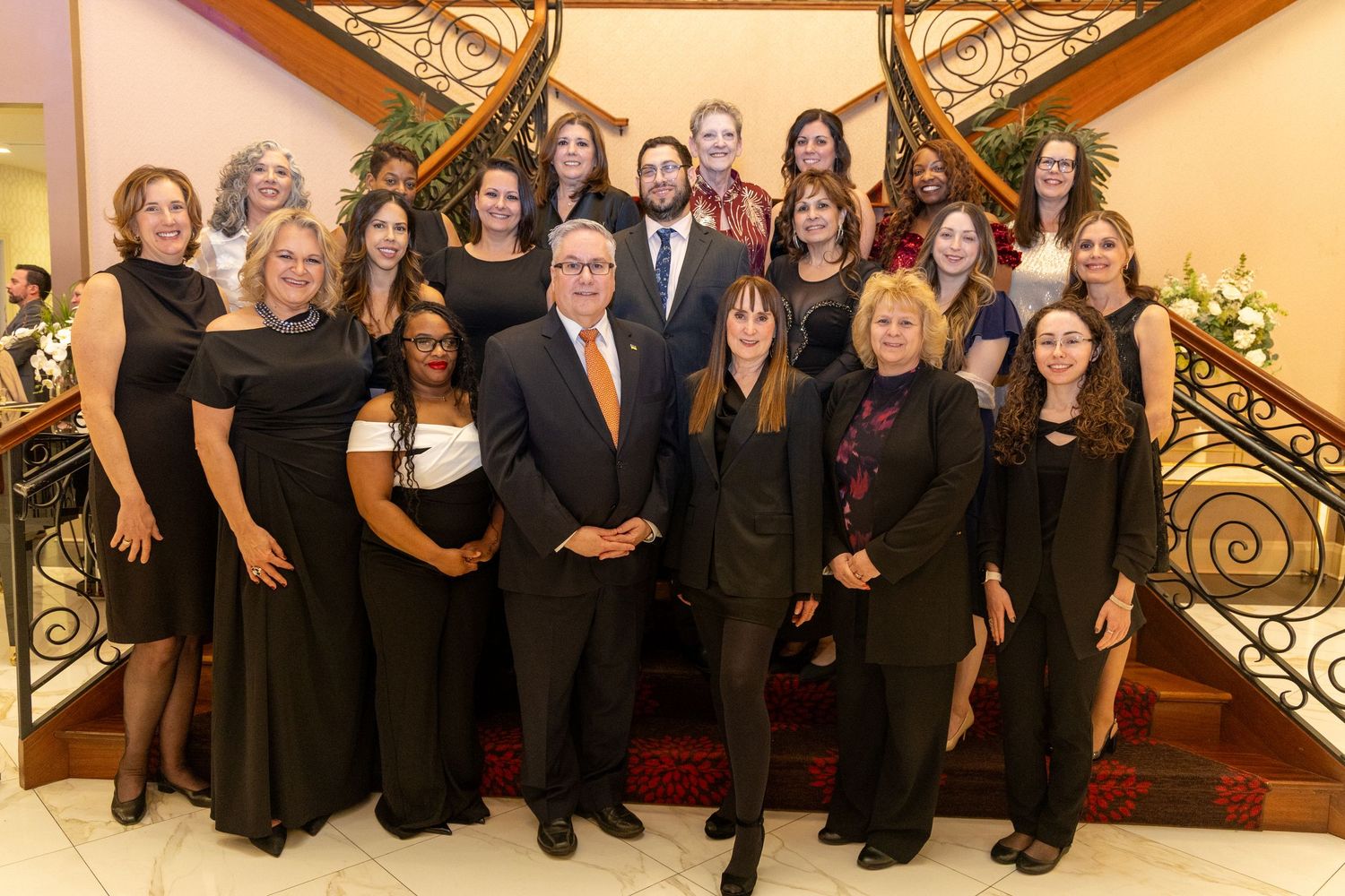 A group of formally dressed people pose together on a grand staircase with ornate gold railings.