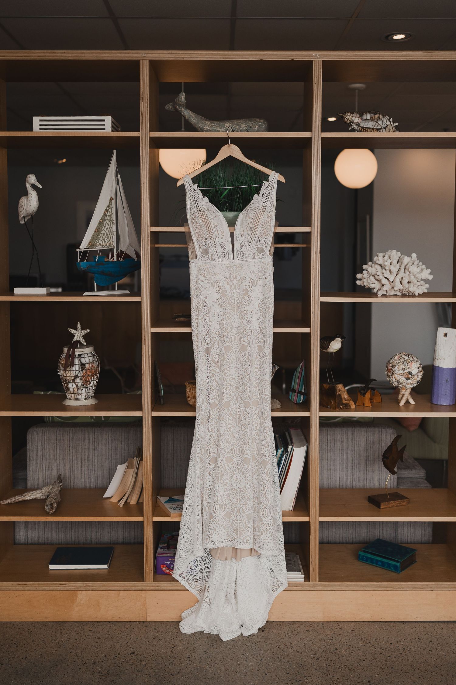 Elegant lace wedding dress hanging on a wooden bookshelf decorated with coastal decor items.