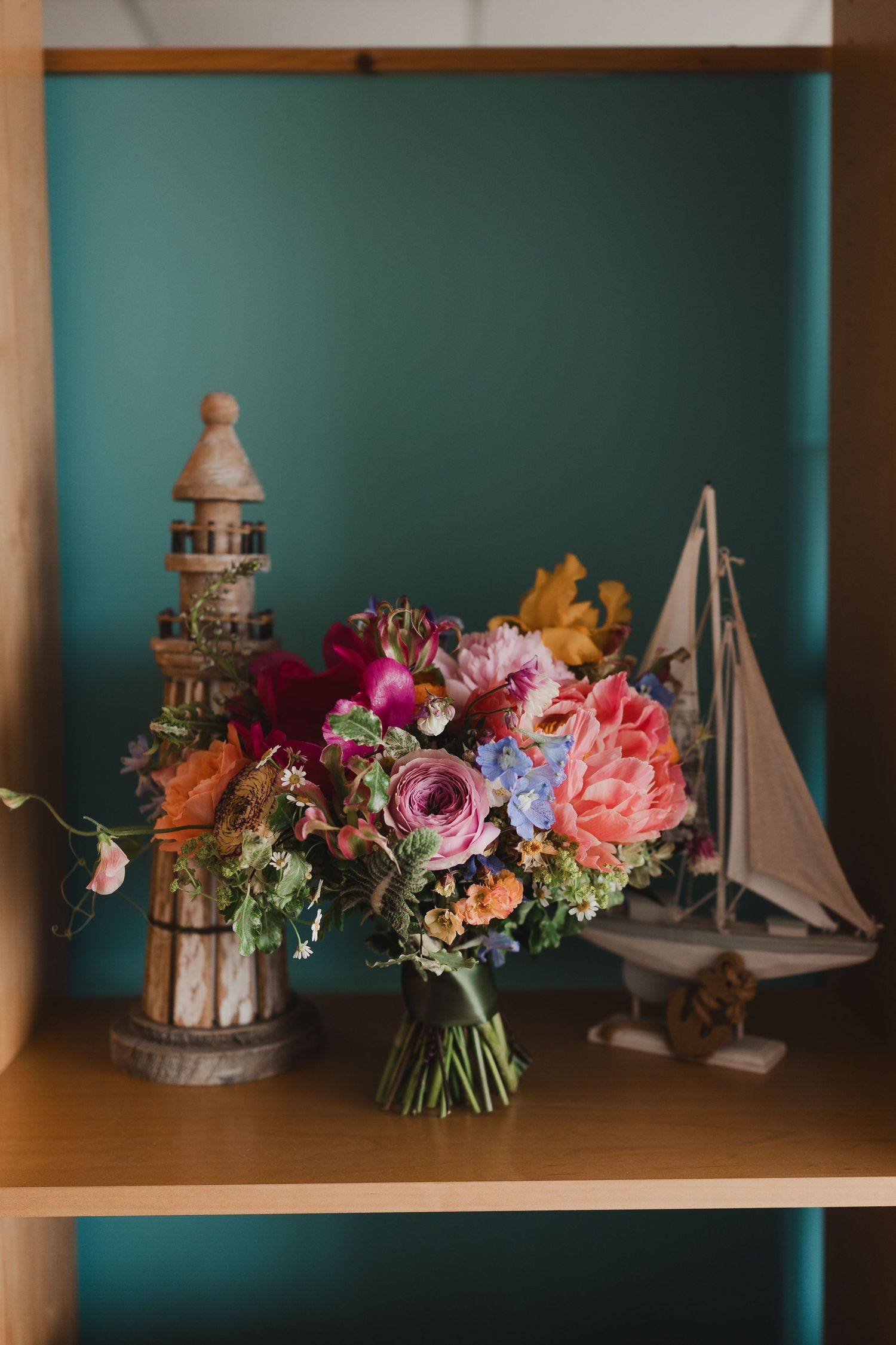 Colorful bridal bouquet with pink peonies and ranunculus on a wooden shelf with nautical lighthouse and sailboat decor.