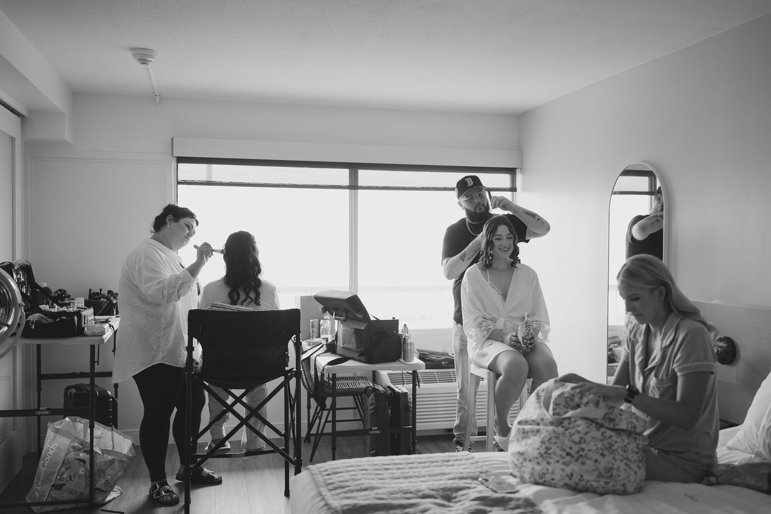 Black and white photo of bridesmaids getting hair and makeup done in a bright hotel room before a wedding.