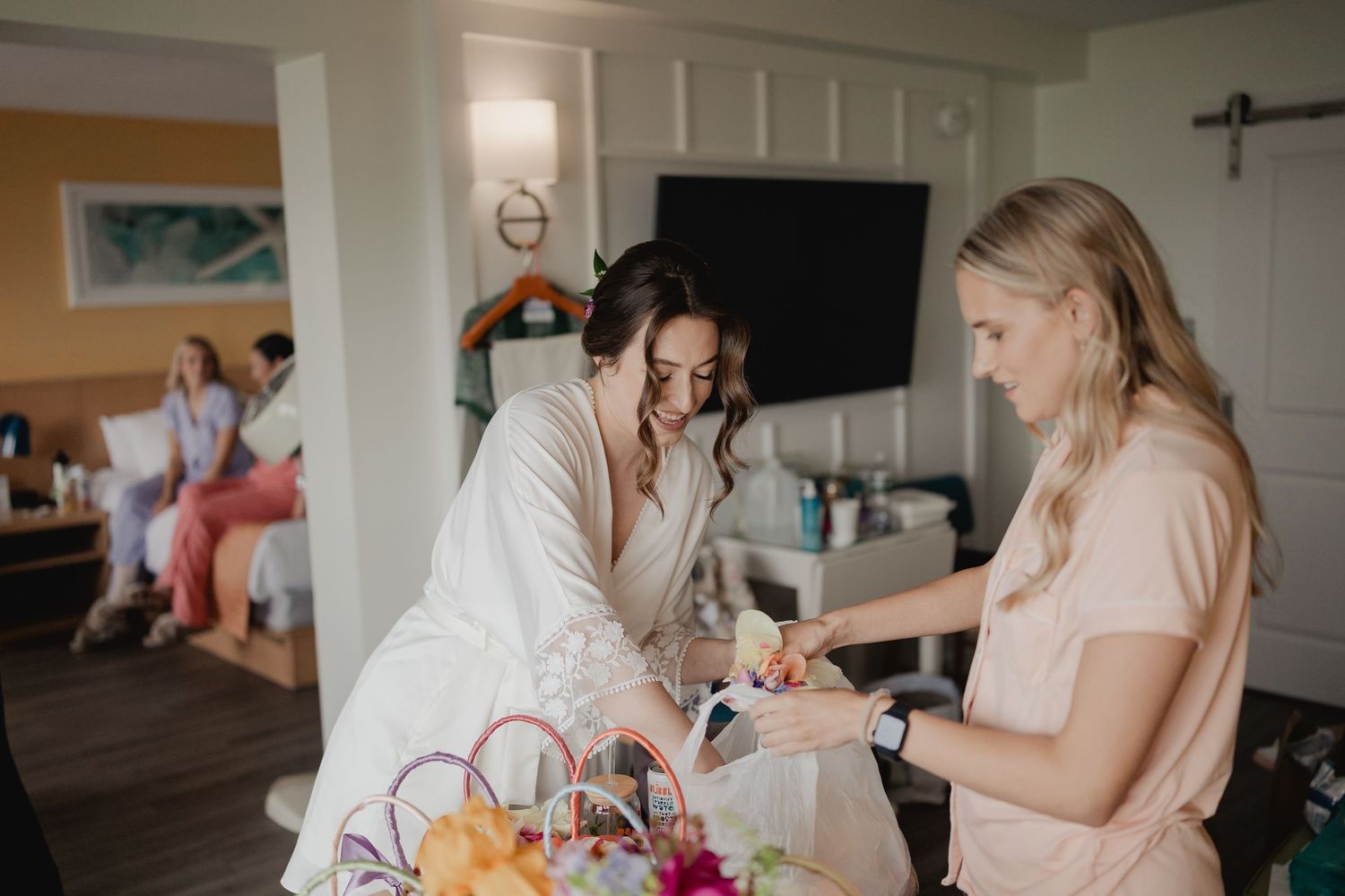 Bride in white robe reviews wedding details with blonde bridesmaid in hotel room before ceremony.