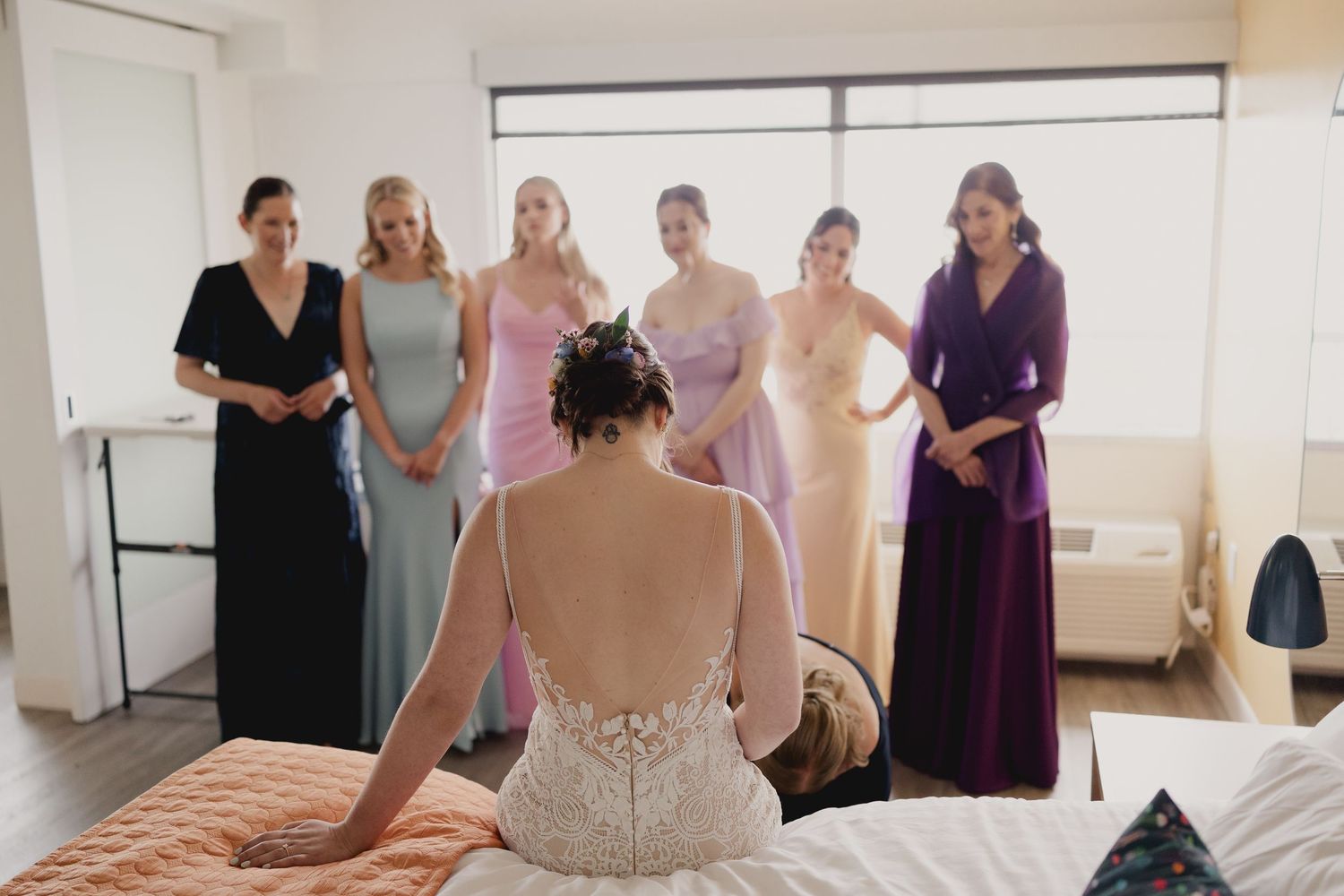 Bride in lace gown sits on bed as bridesmaids in colorful dresses look on during wedding preparations.