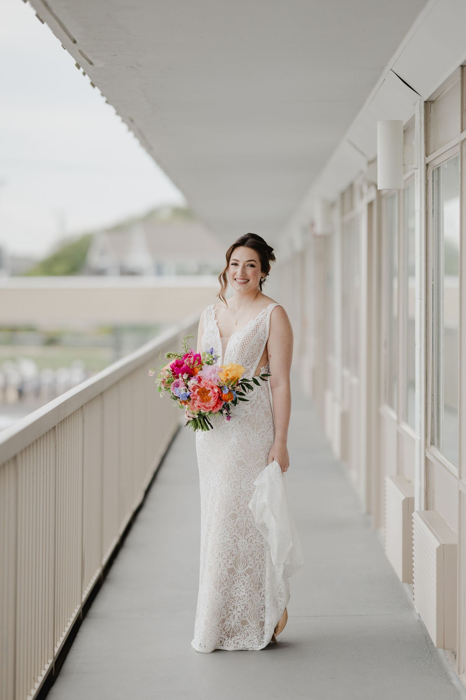 Smiling bride in lace gown holding colorful bouquet walks along outdoor corridor on her wedding day.