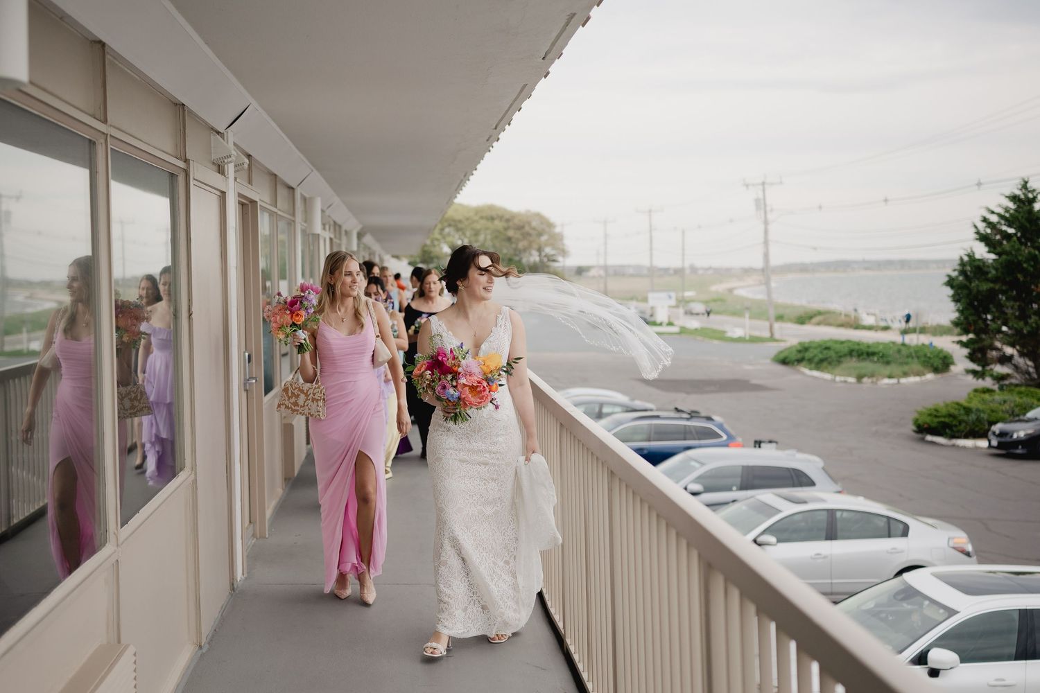 Bride in white lace gown walks with bridesmaids in pink dresses along a waterfront venue corridor.