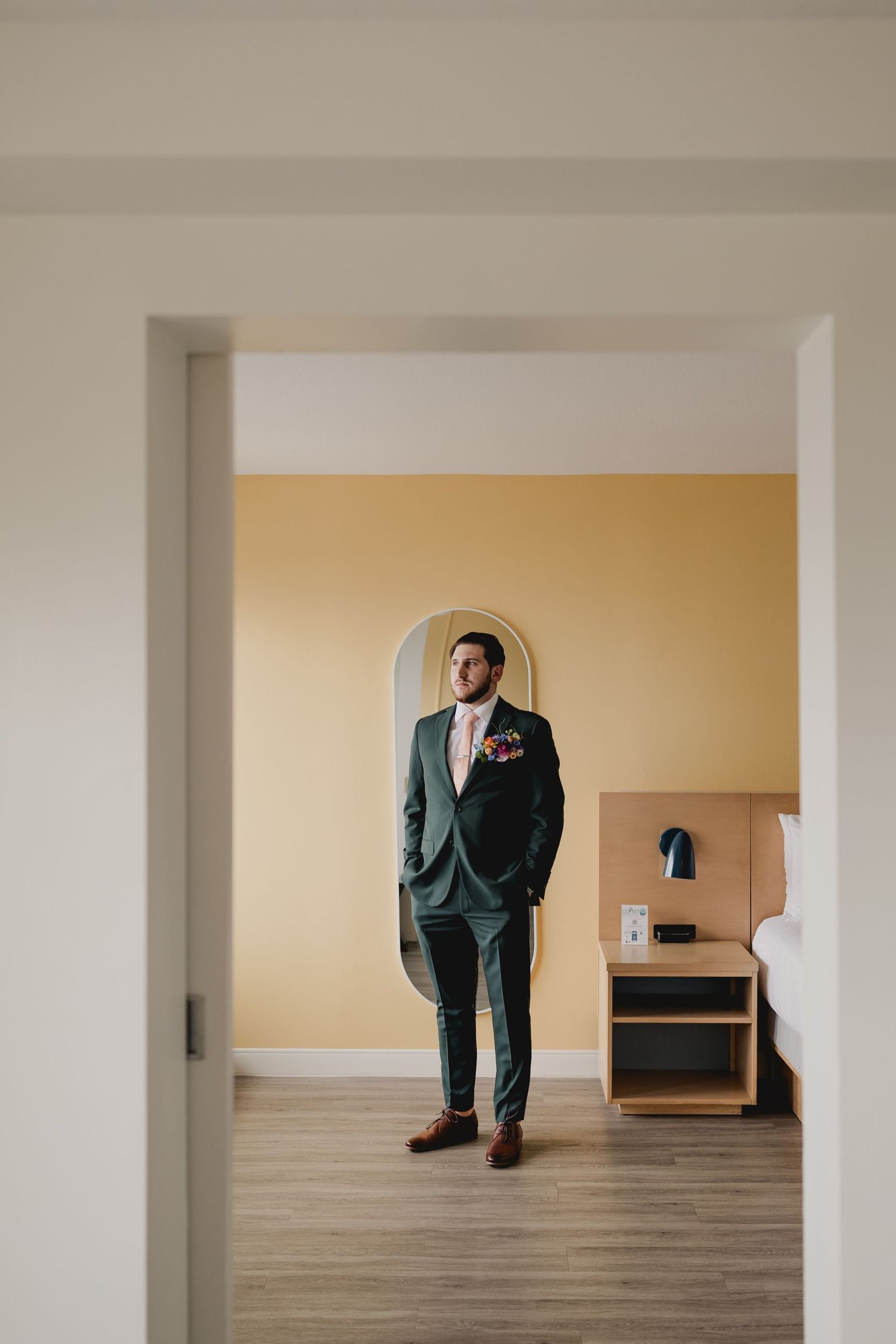 Groom in dark green suit with floral boutonniere stands confidently in a hotel room with yellow walls.