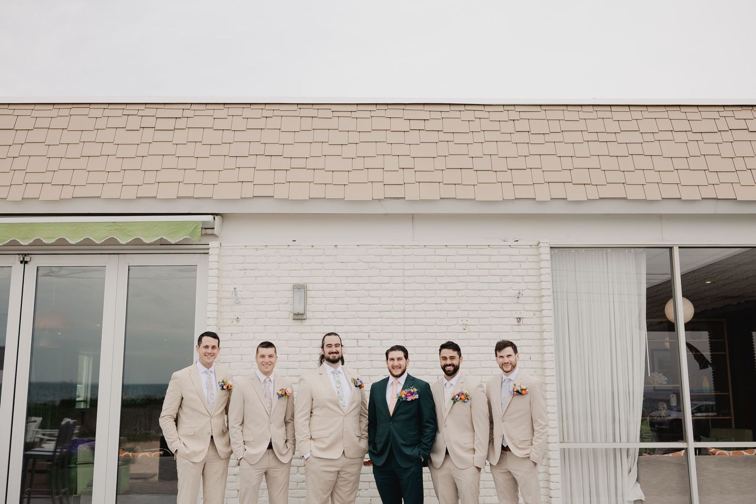 Groom in dark teal suit poses with five groomsmen in tan suits outside a white brick building.