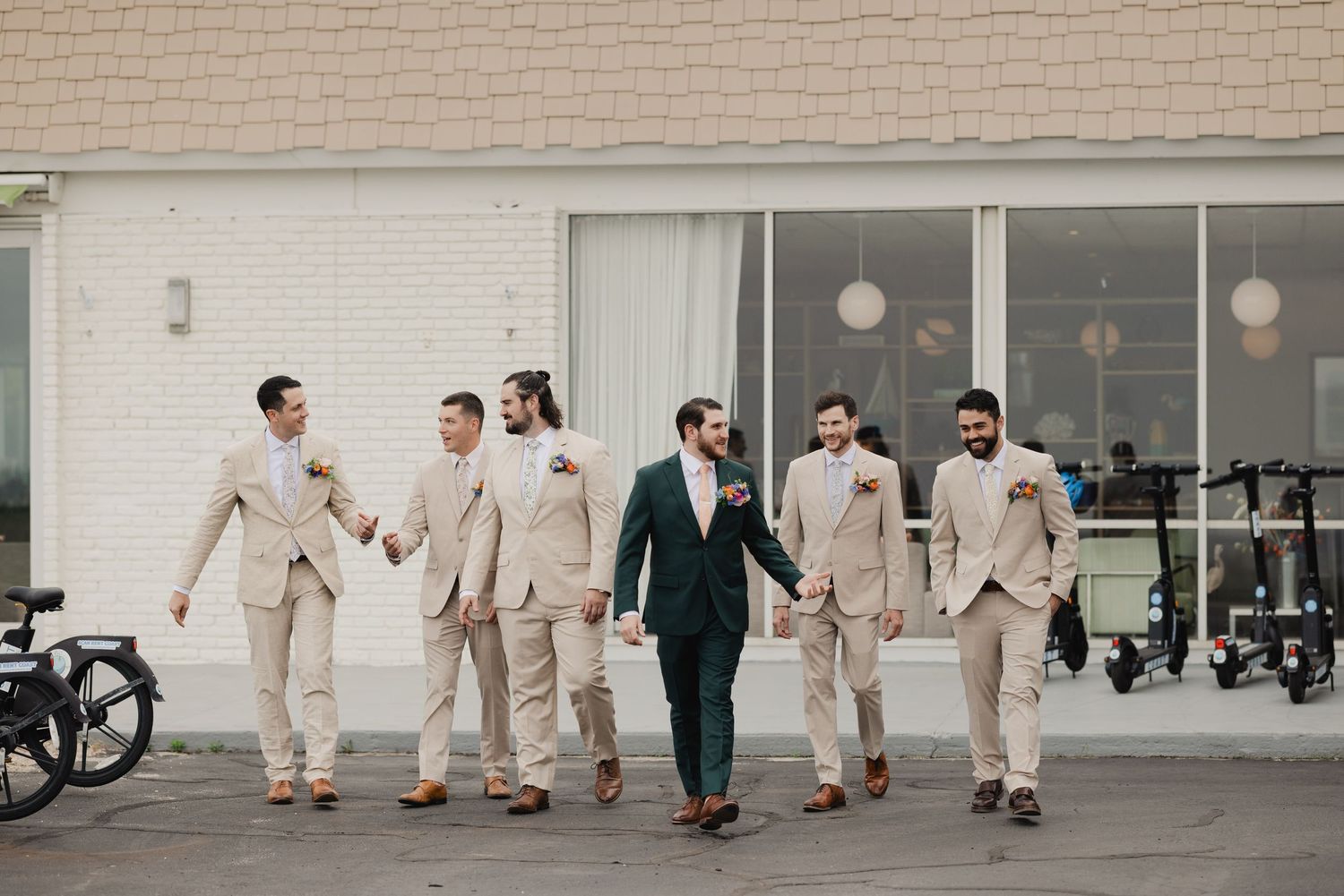 Groom in navy suit walks with groomsmen in beige suits outside a modern white brick venue.