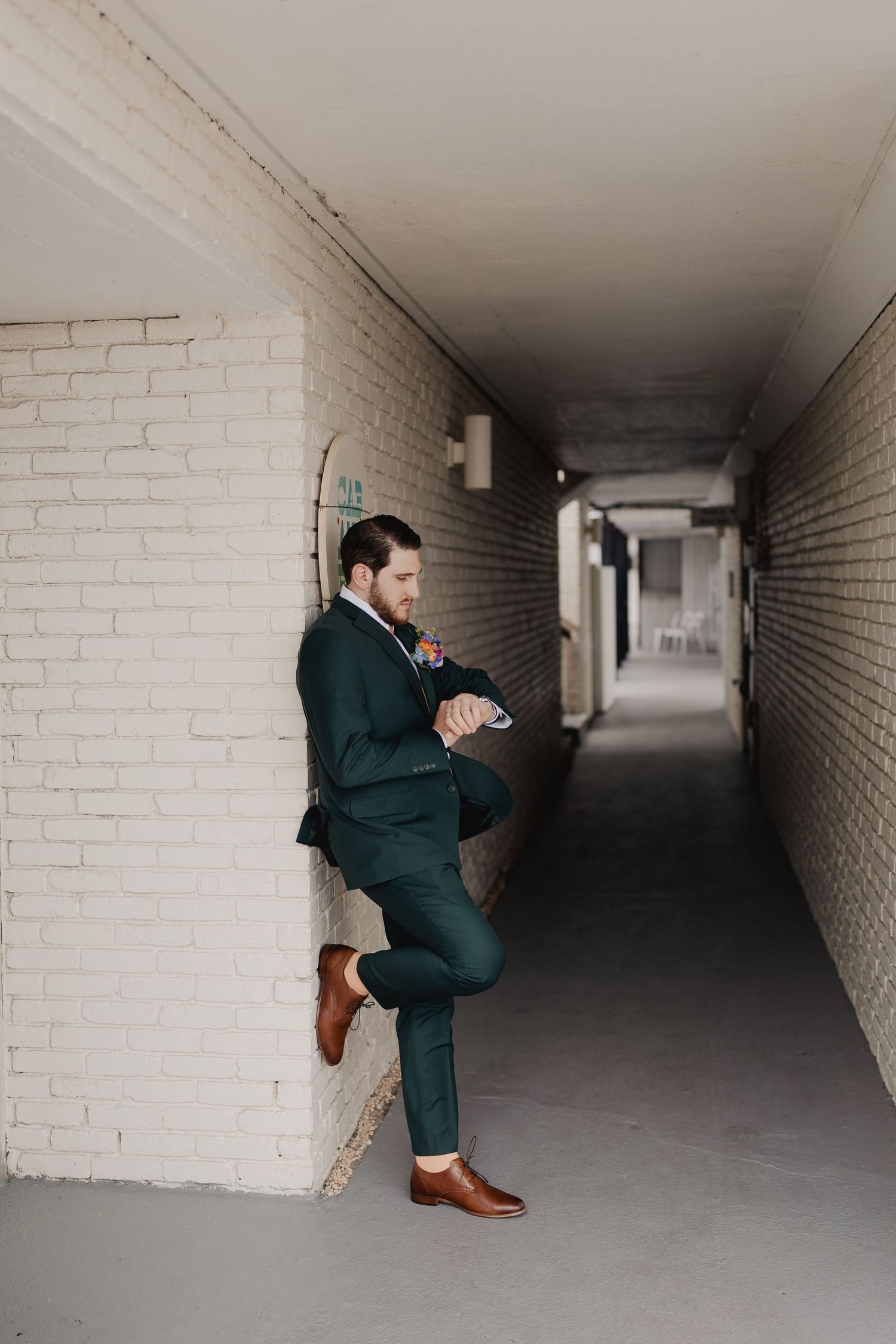 Dapper groom in dark green suit leaning against white brick wall in corridor.