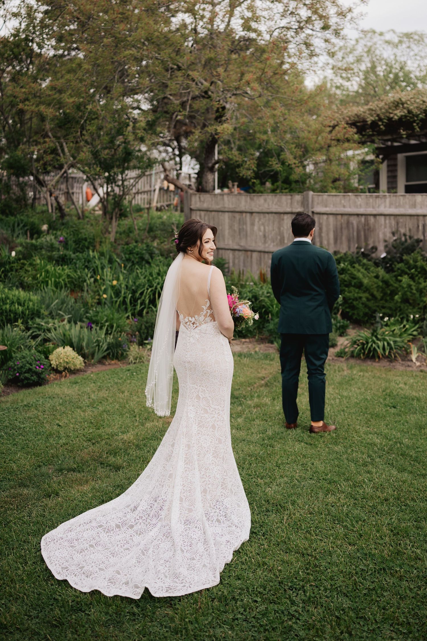 Bride in lace gown with veil approaches groom for first look in lush garden.