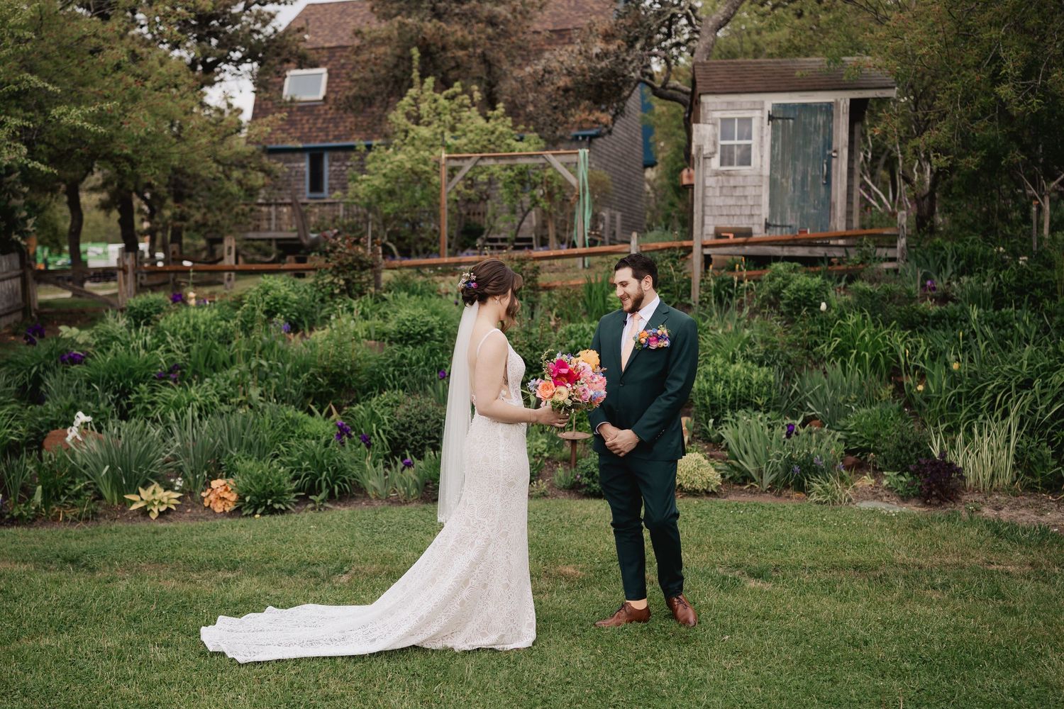 Bride and groom share first look moment in garden with rustic building backdrop.