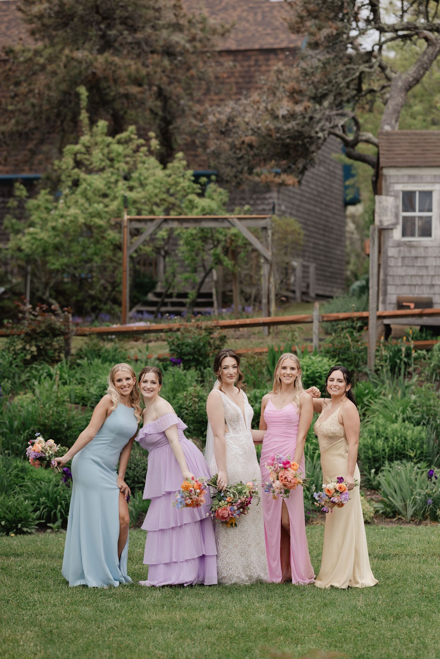Five bridesmaids in pastel mismatched gowns holding colorful bouquets pose outdoors at a garden wedding venue.