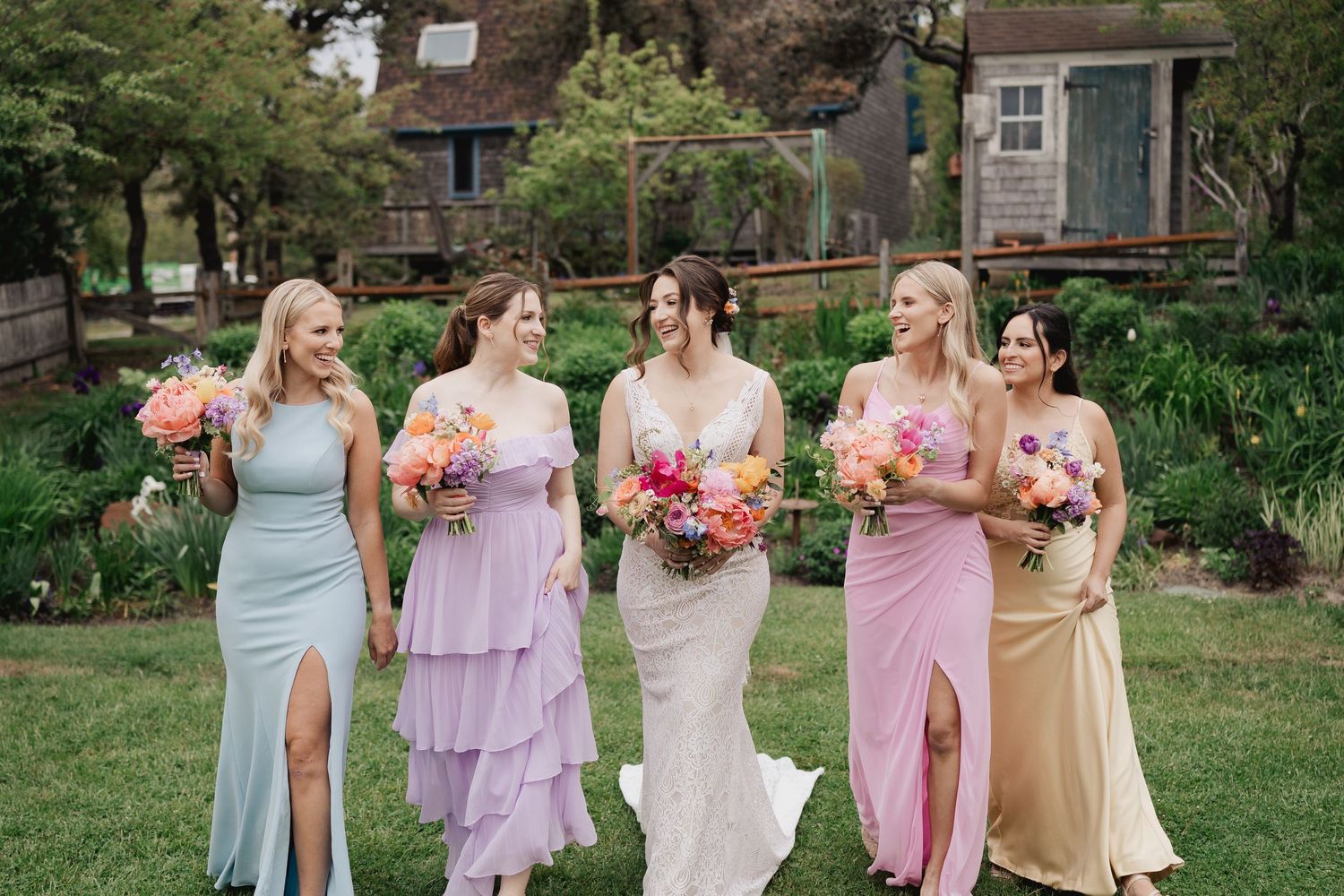 Bride walks with four bridesmaids in colorful mismatched dresses holding vibrant bouquets in a garden setting.