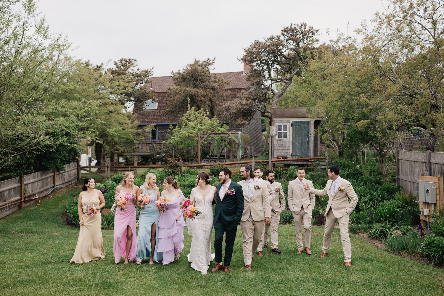 Wedding party in colorful dresses and tan suits walks through lush garden outside rustic venue.