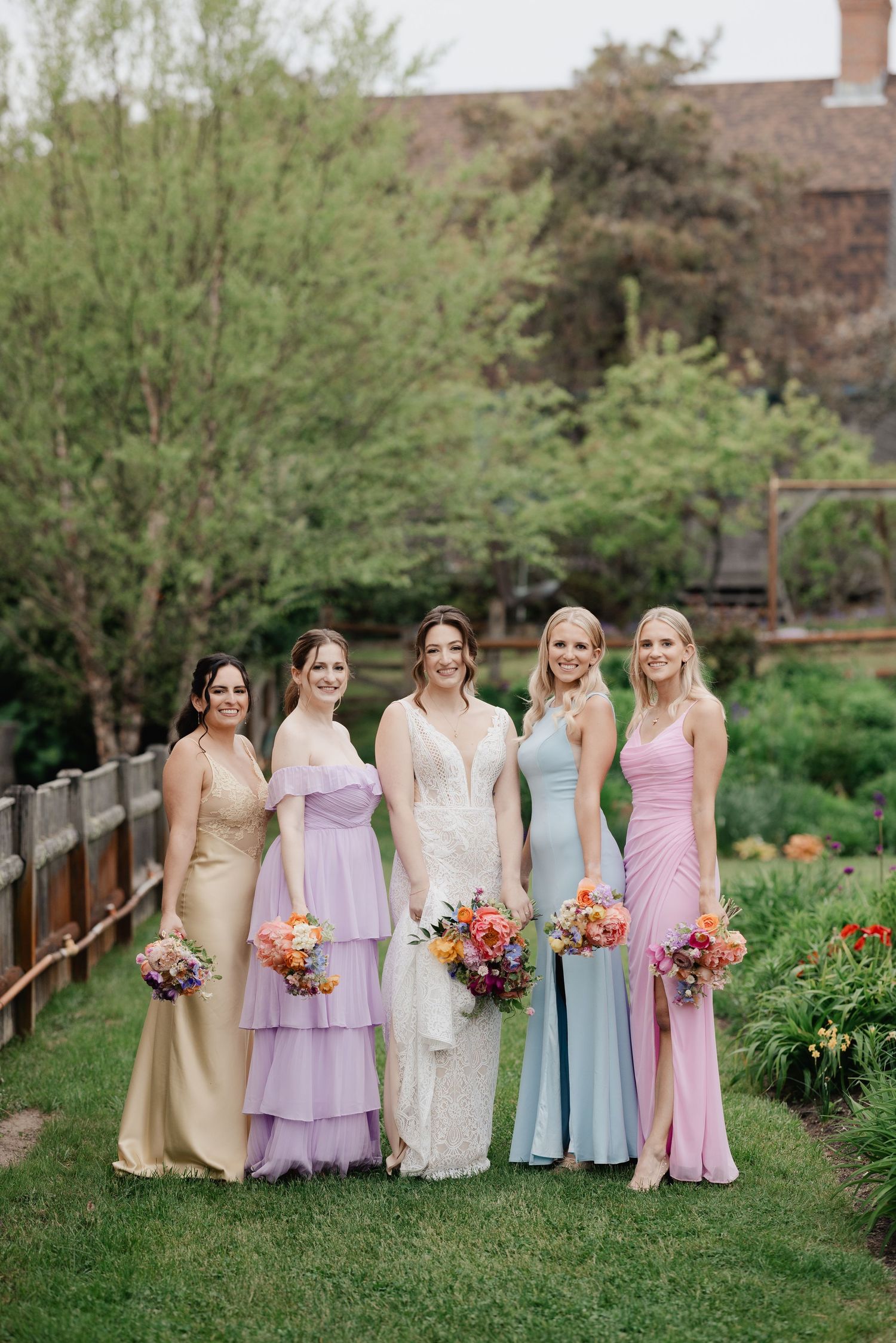 Bride with four bridesmaids in mismatched pastel gowns holding colorful bouquets outdoors.