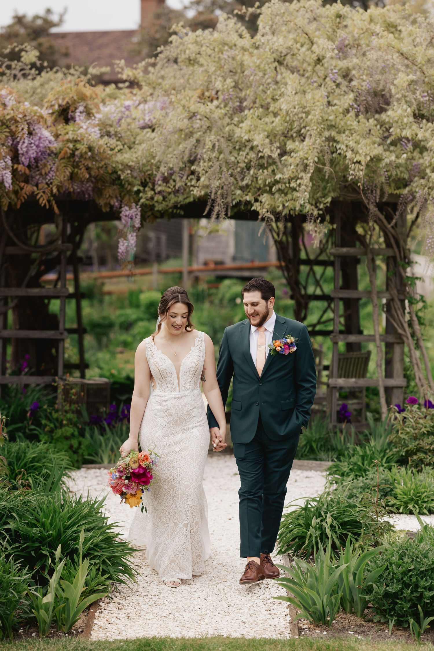 A bride and groom walk hand in hand through a lush garden pergola, she holds a vibrant bouquet.