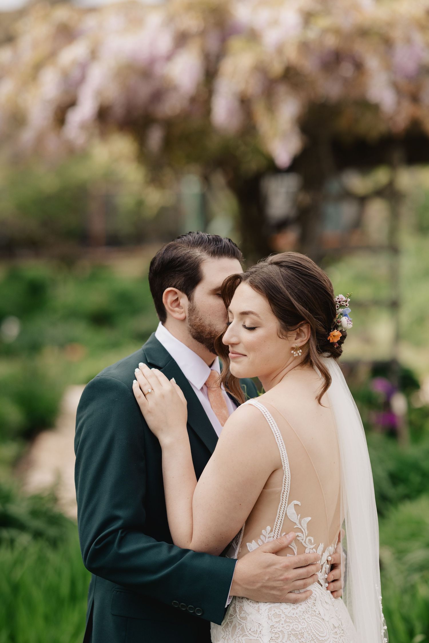 A groom in a dark green suit kisses his bride wearing a lace backless gown under blooming wisteria in a garden.