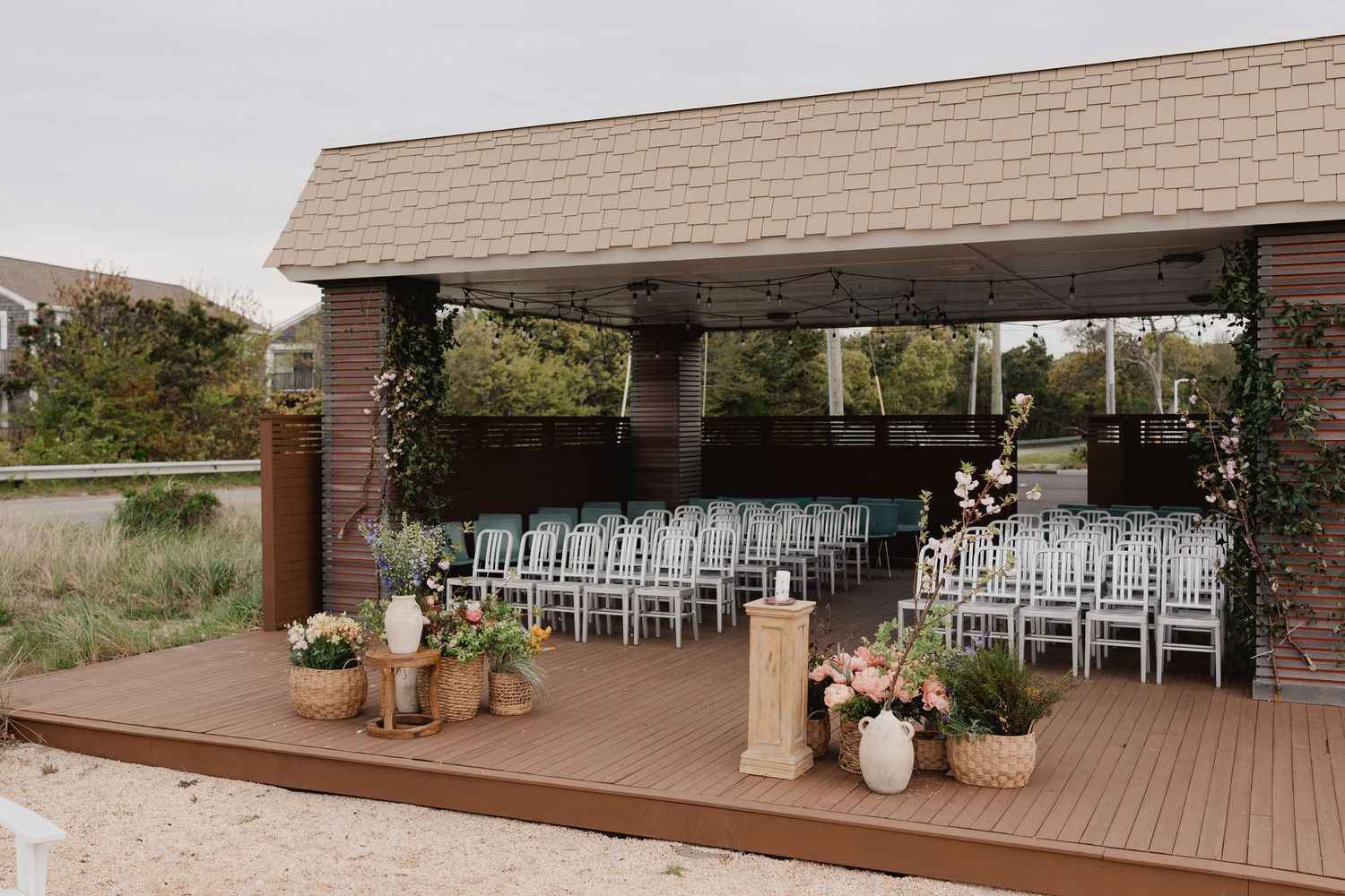 Elegant outdoor wedding ceremony setup with white chairs under a wooden pavilion decorated with floral arrangements.