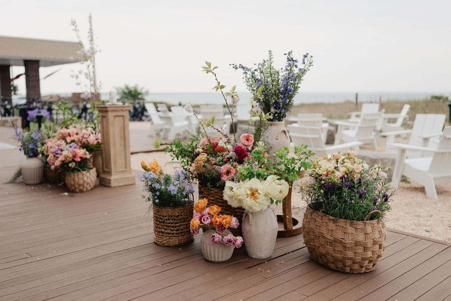 Assorted floral arrangements in wicker baskets and vases displayed on a wooden deck at a beach wedding.