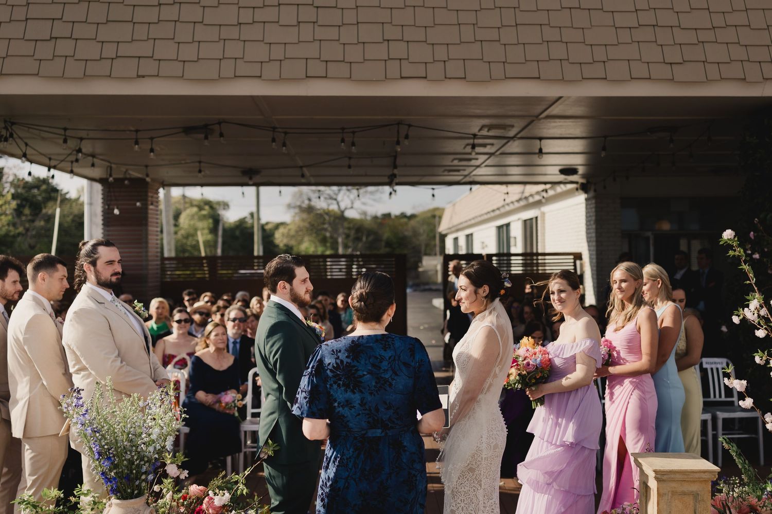 Wedding guests congratulate the newlyweds during an outdoor ceremony under a covered venue with string lights.