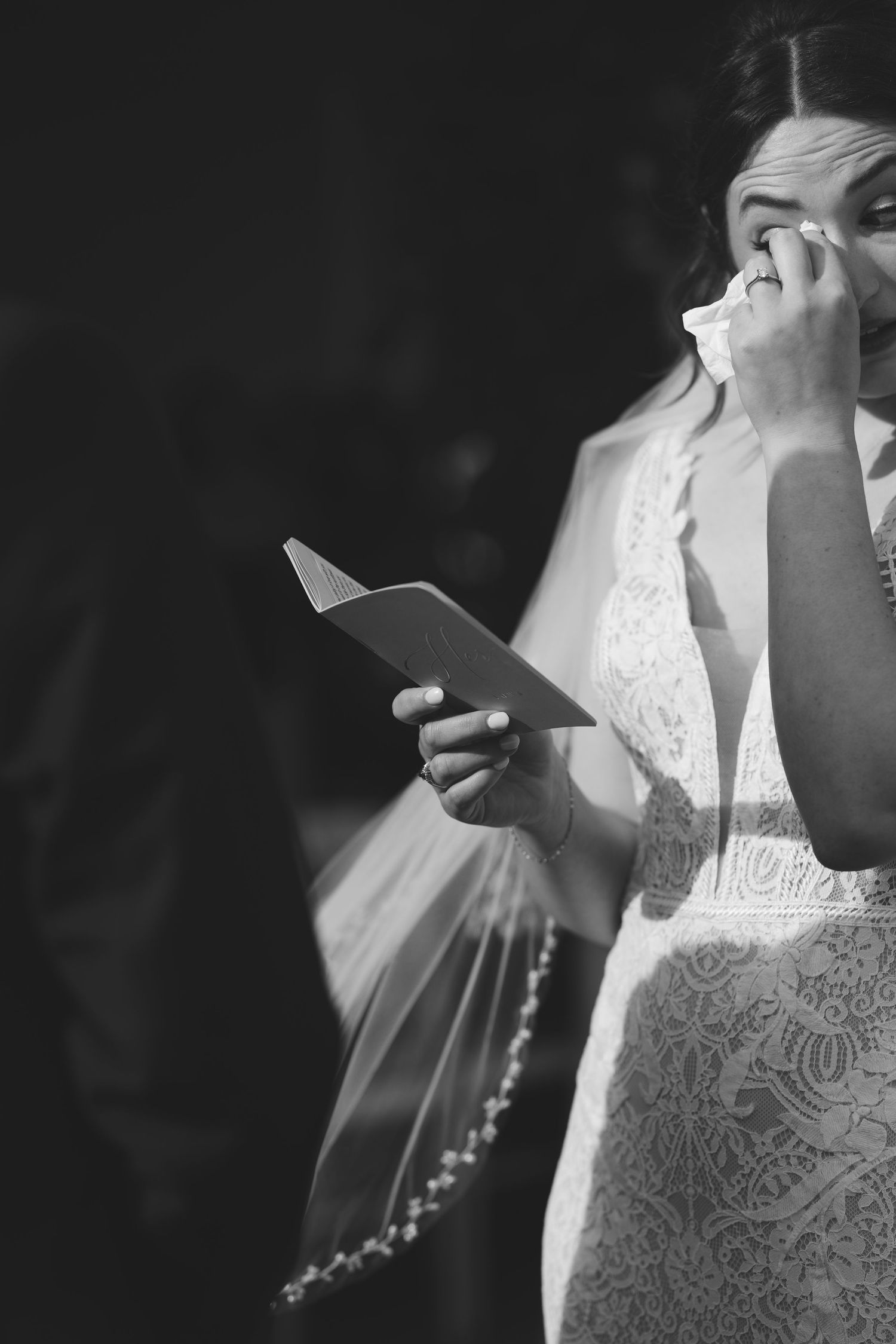 Emotional bride in lace gown wipes tears while reading vows during wedding ceremony, black and white.