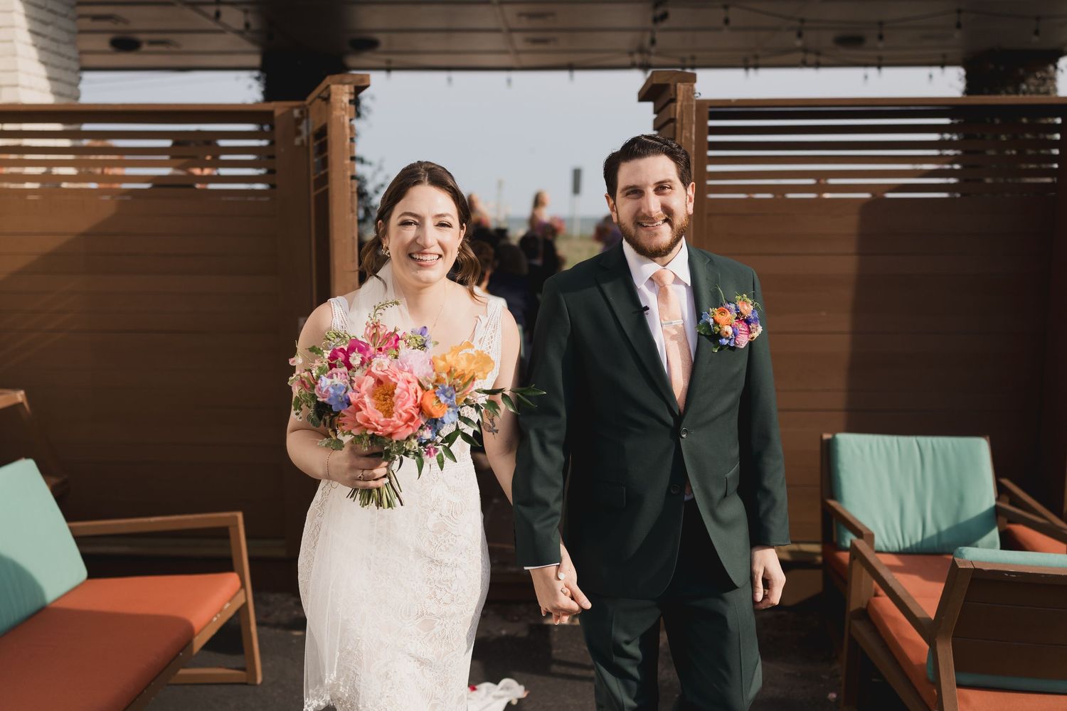 Newlyweds walk hand in hand smiling with colorful bridal bouquet on rooftop venue.