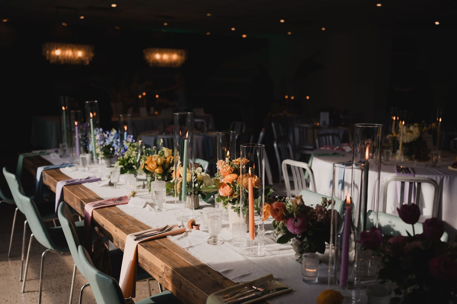 Elegant wedding reception table with wooden surface, colorful candles, and floral centerpieces in a dimly lit venue.