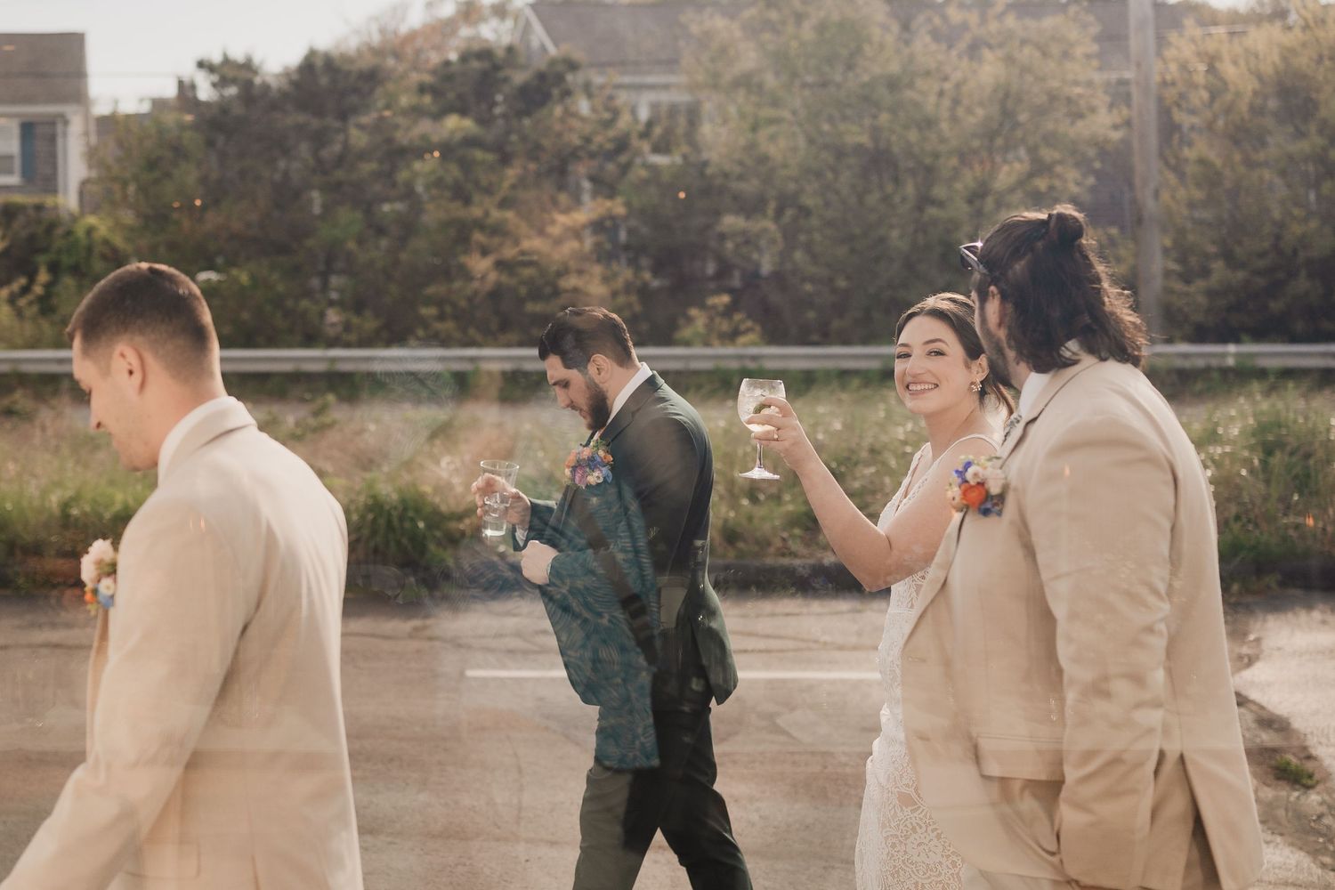 Bride and groom toast champagne with groomsmen while walking outdoors in golden sunlight.