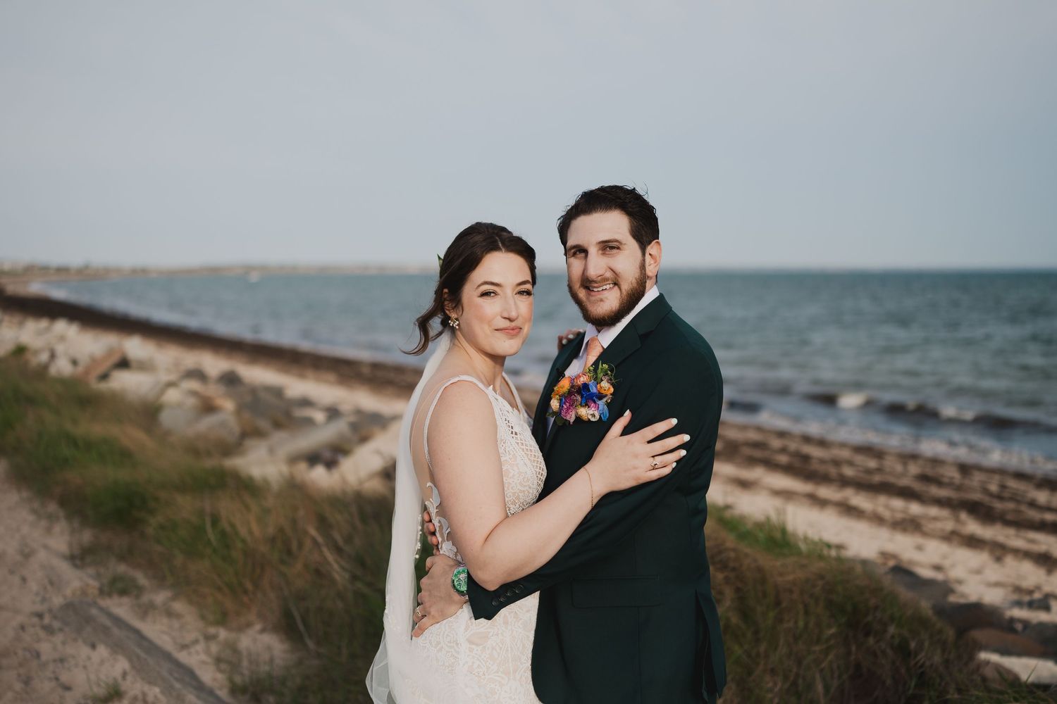 Bride and groom embrace near rocky shoreline with ocean backdrop at their beach wedding.