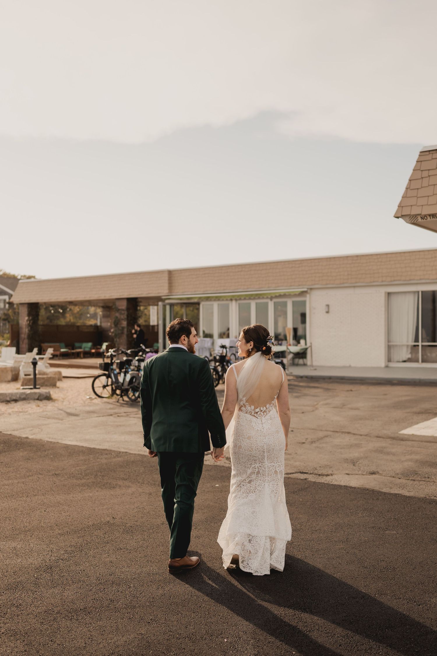 Bride in lace backless gown and groom in dark suit walk hand-in-hand toward a modern venue building.