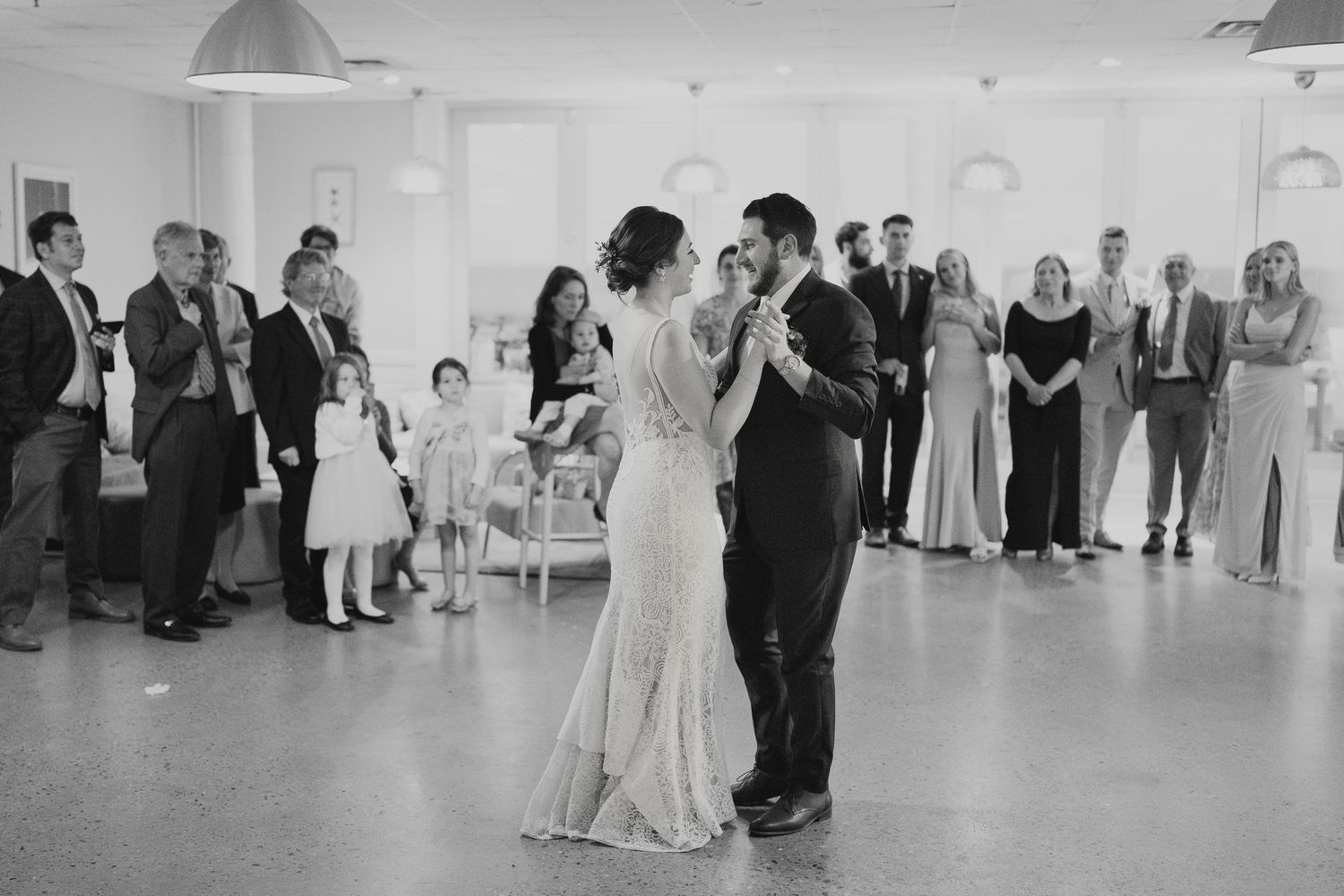 Bride and groom share first dance surrounded by guests in elegant white reception hall, black and white.