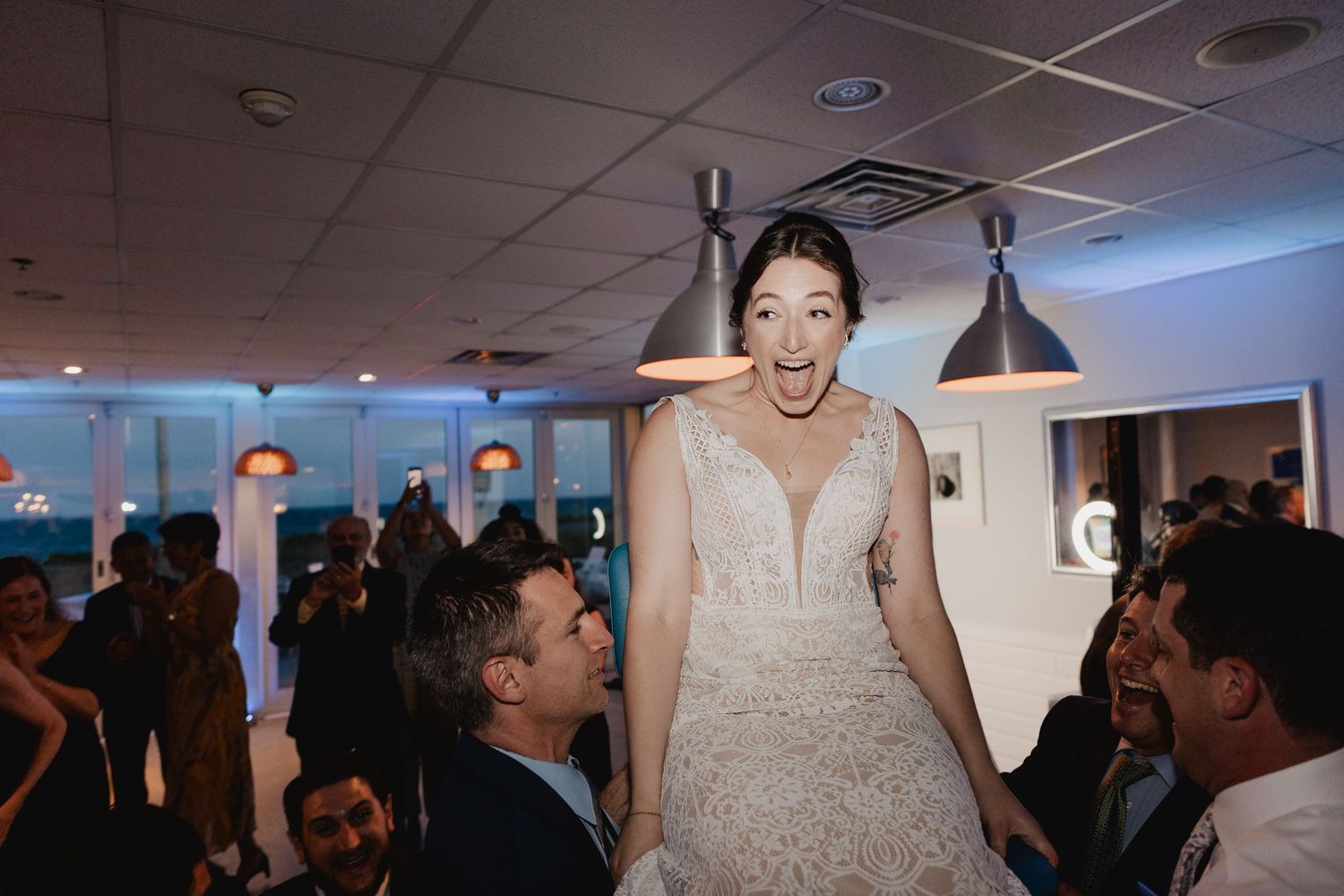 Joyful bride in lace gown lifted by guests on the dance floor during wedding reception celebration.