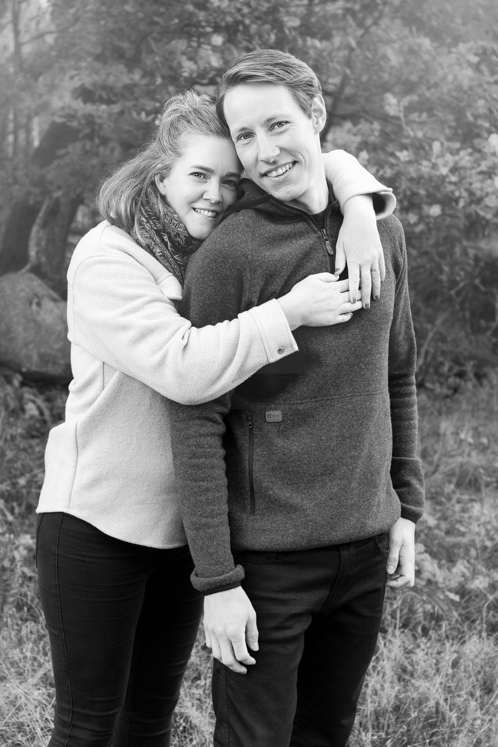 Black and white photo of a smiling couple embracing outdoors, woman resting head on mans shoulder among trees.