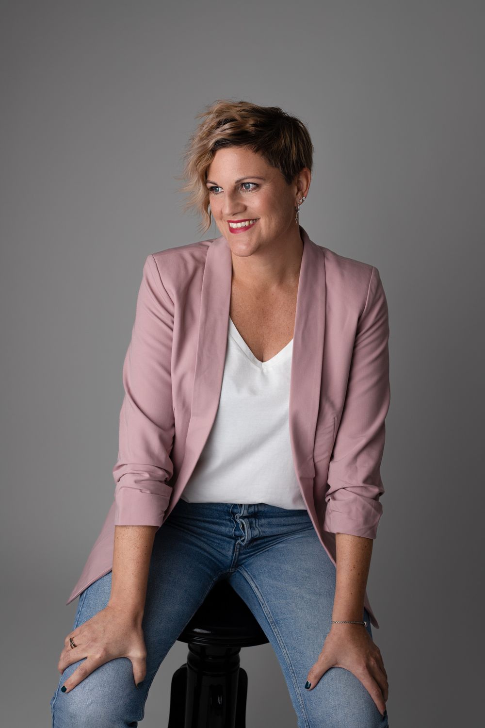 Woman in pink blazer and jeans sits smiling on stool against gray background.