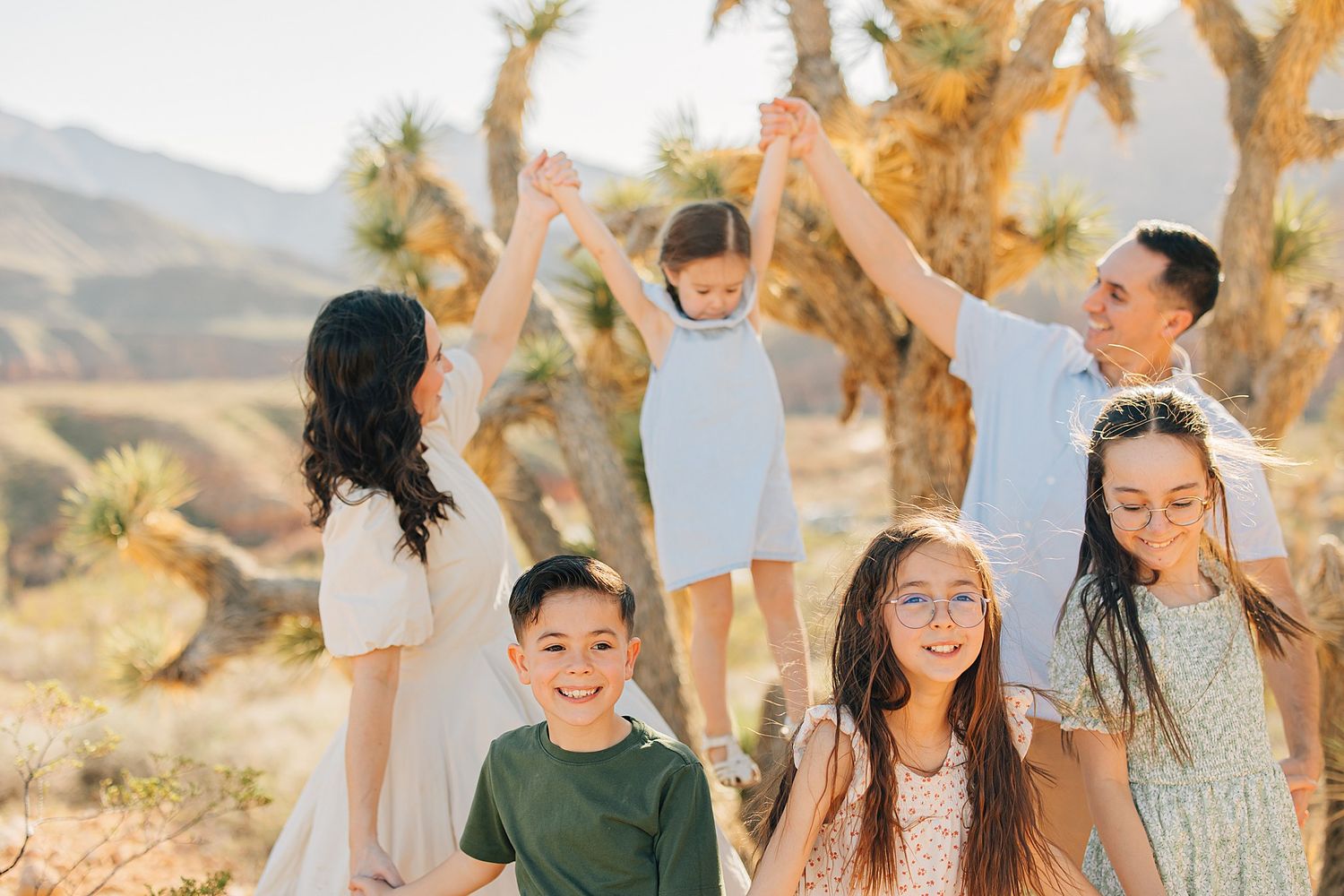 Family of five posing outdoors in a desert landscape with Joshua trees, parents lifting toddler while children smile.