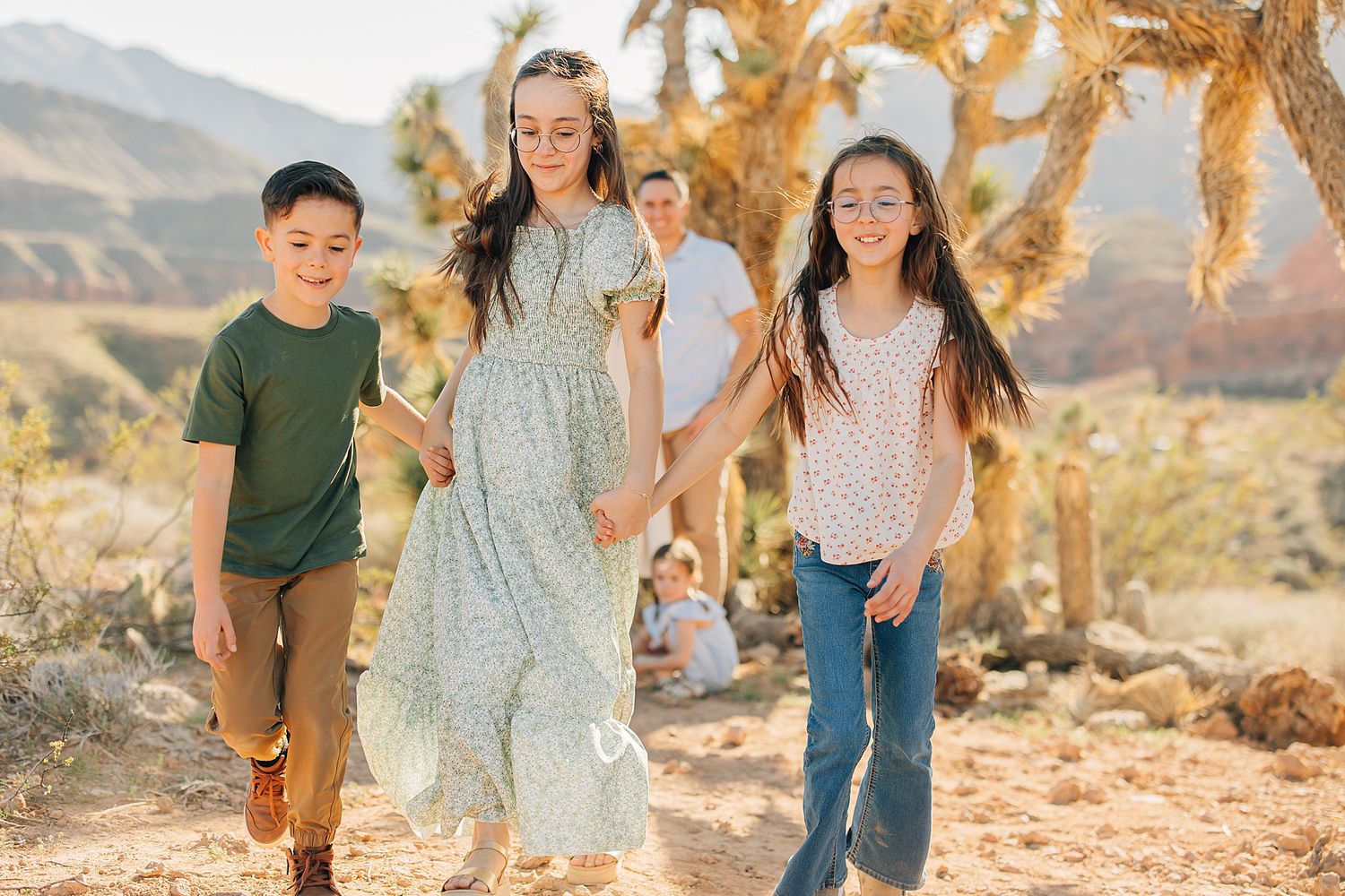 Family walking hand-in-hand through a desert landscape with Joshua trees and mountains in the background on a sunny day.