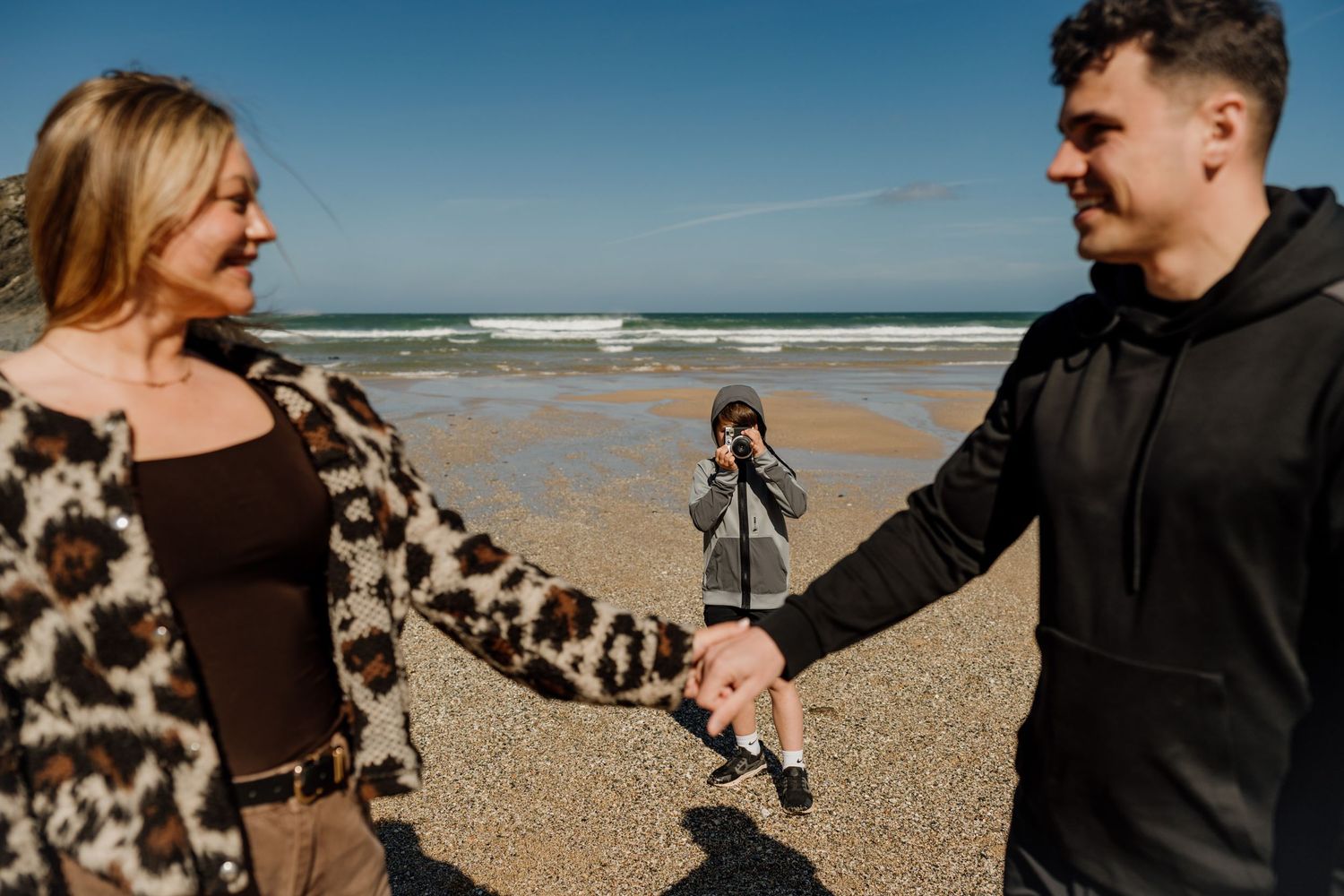 Couple holding hands on a sunny beach with a child standing between them near the ocean shoreline.