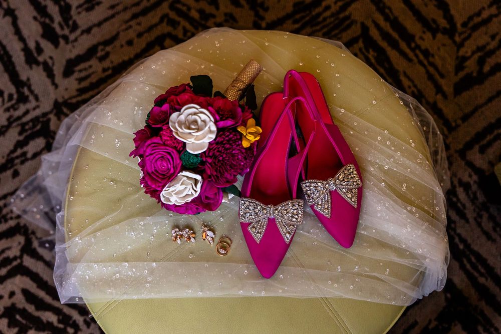 Fuchsia bridal heels with rhinestone bows beside a vibrant pink floral bouquet and gold earrings on a white chair.