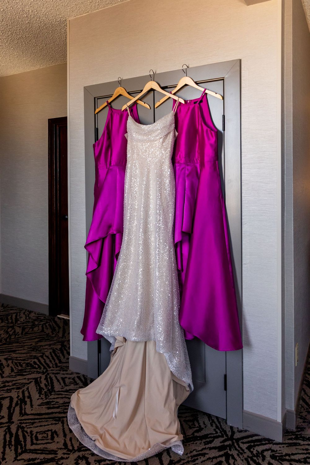 White sequin wedding gown flanked by magenta bridesmaid dresses hanging on wooden hangers in the Double Tree in downtown Omaha.