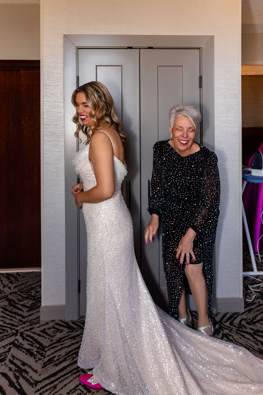 Bride in white sequin gown with train laughs with mom in black dress in the Double Tree in downtown Omaha.