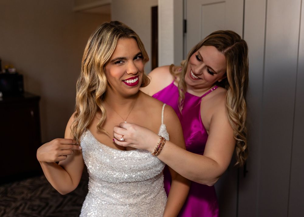 Bride in white sequin gown gets help from bridesmaid in magenta dress, both smiling during wedding preparations at the Double Tree in downto