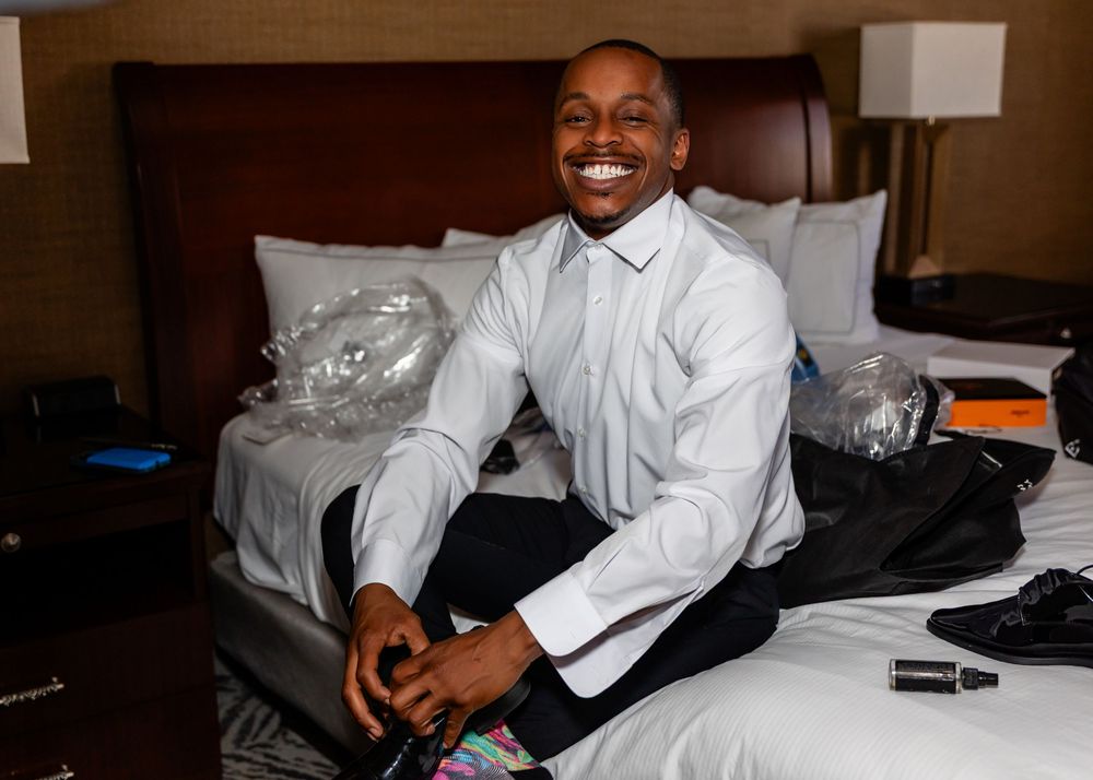 Groom in white dress shirt sitting on hotel bed with formal accessories nearby at the Double Tree in downtown Omaha.