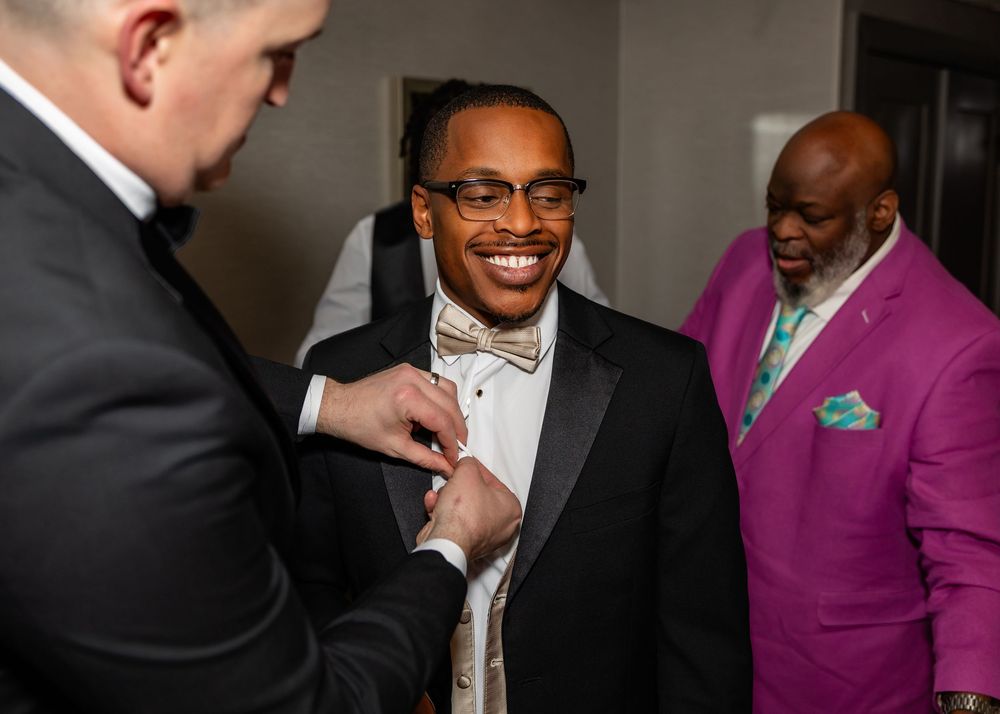 Groom in tuxedo smiling while getting boutonniere pinned, with man in purple suit standing nearby in the Double Tree in downtown Omaha.