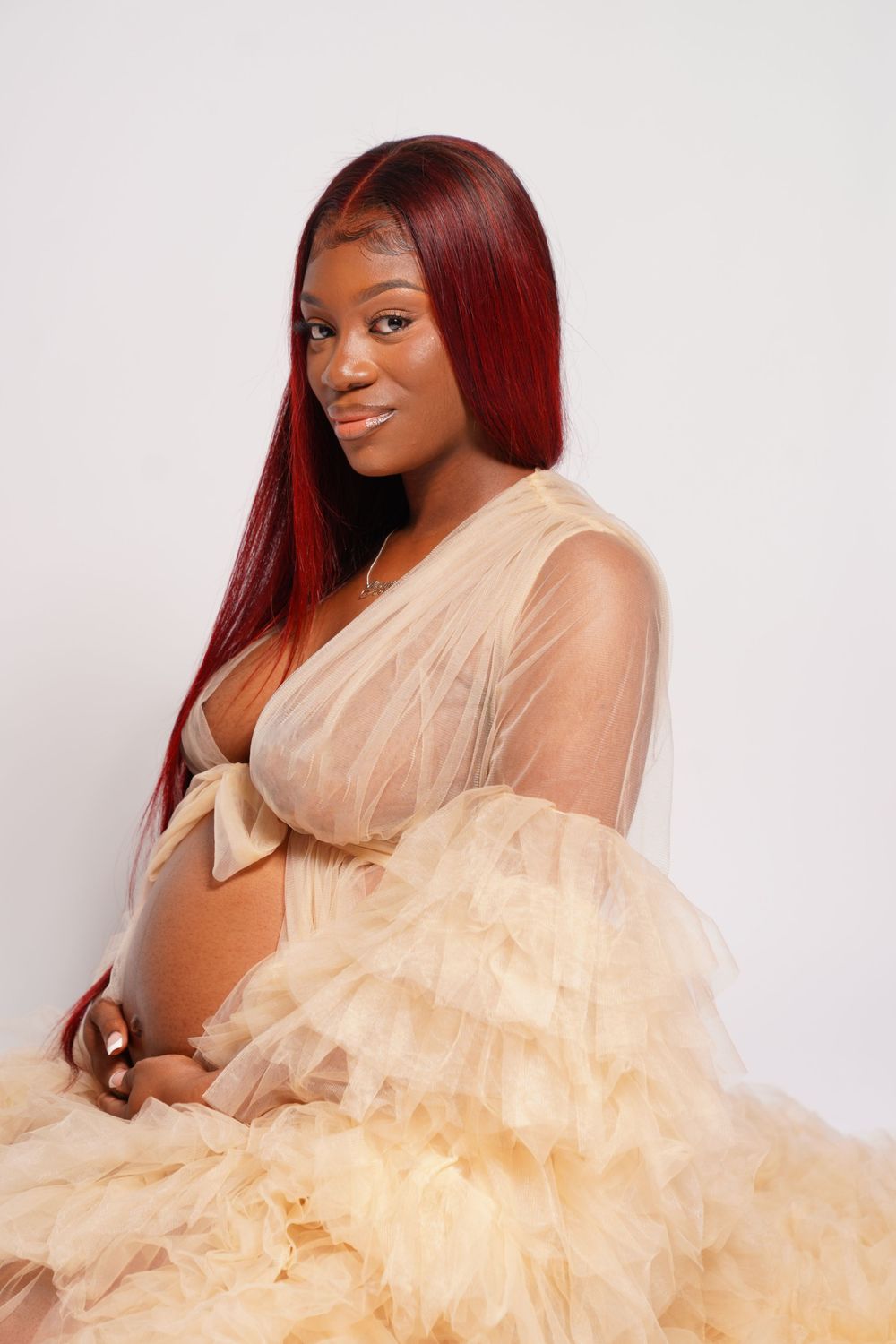 Pregnant woman in sheer champagne tulle robe with red hair poses gracefully against white background.
