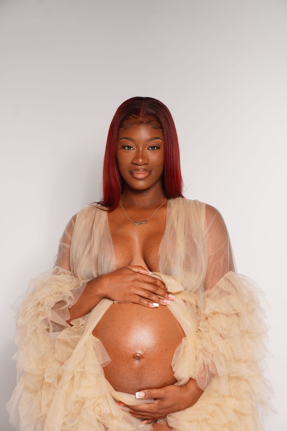 Pregnant Black woman with red hair wearing sheer beige robe, cradling baby bump, smiling at camera against white background.